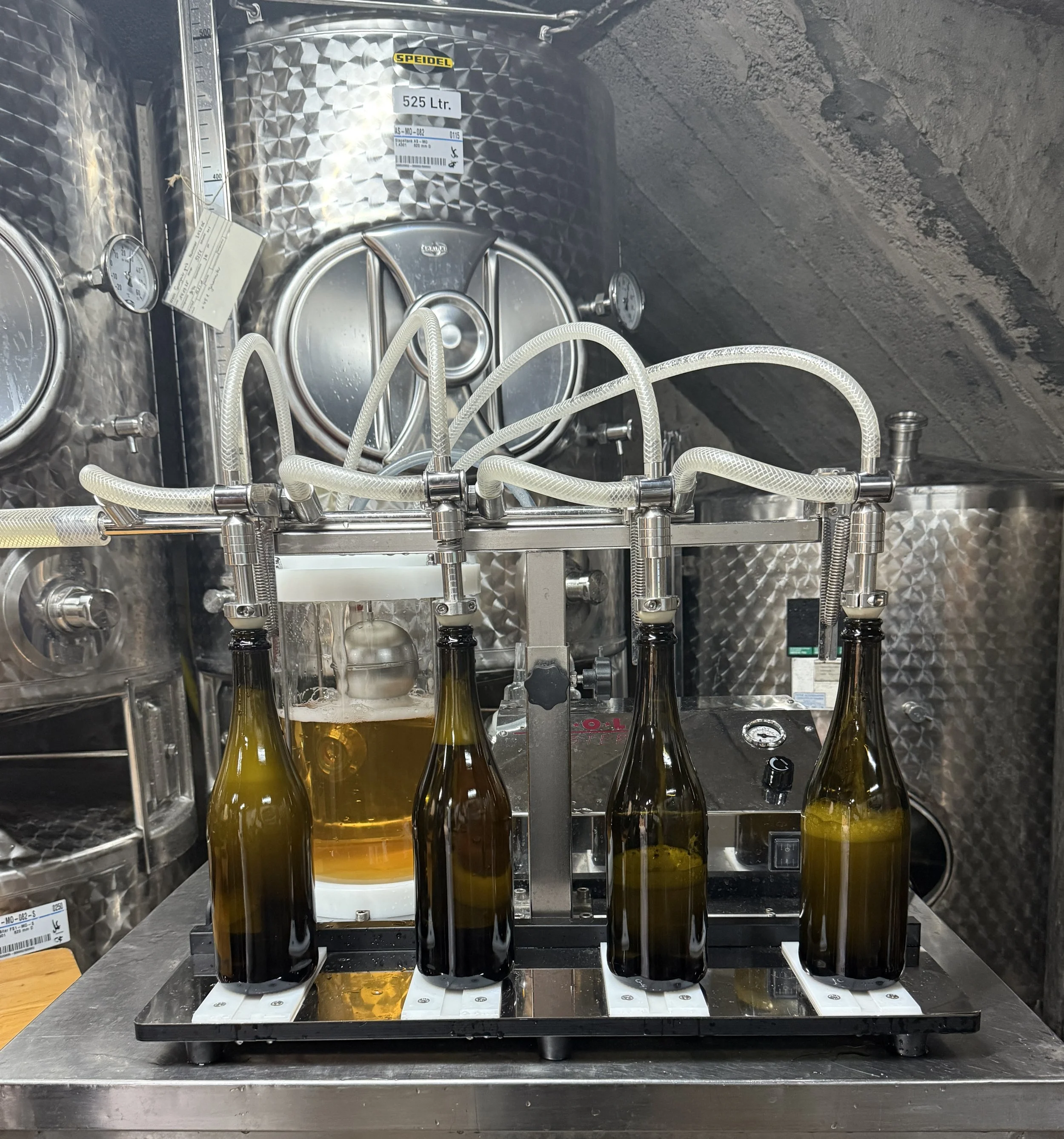 Five glass bottles filled with yellow liquid are positioned in a machine for filling or processing. The machine has tubes connected to the bottles and gauges measuring pressure or flow, with a metallic, textured background.