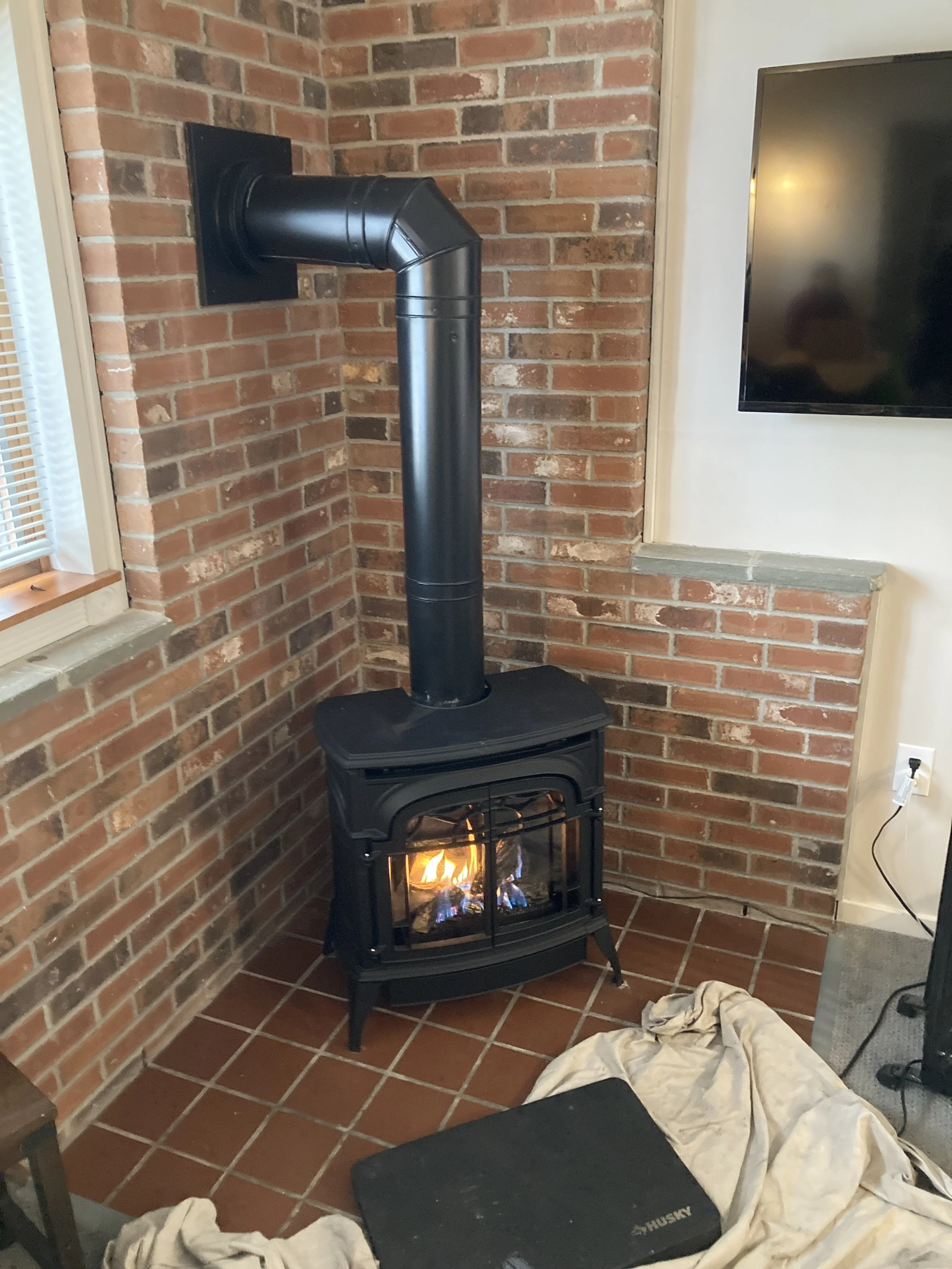 A black gas stove with a glass door showing a fire inside, connected to a black stovepipe venting through a brick wall in a cozy living space. installation just completed by Silas Miller Stove & Chimney