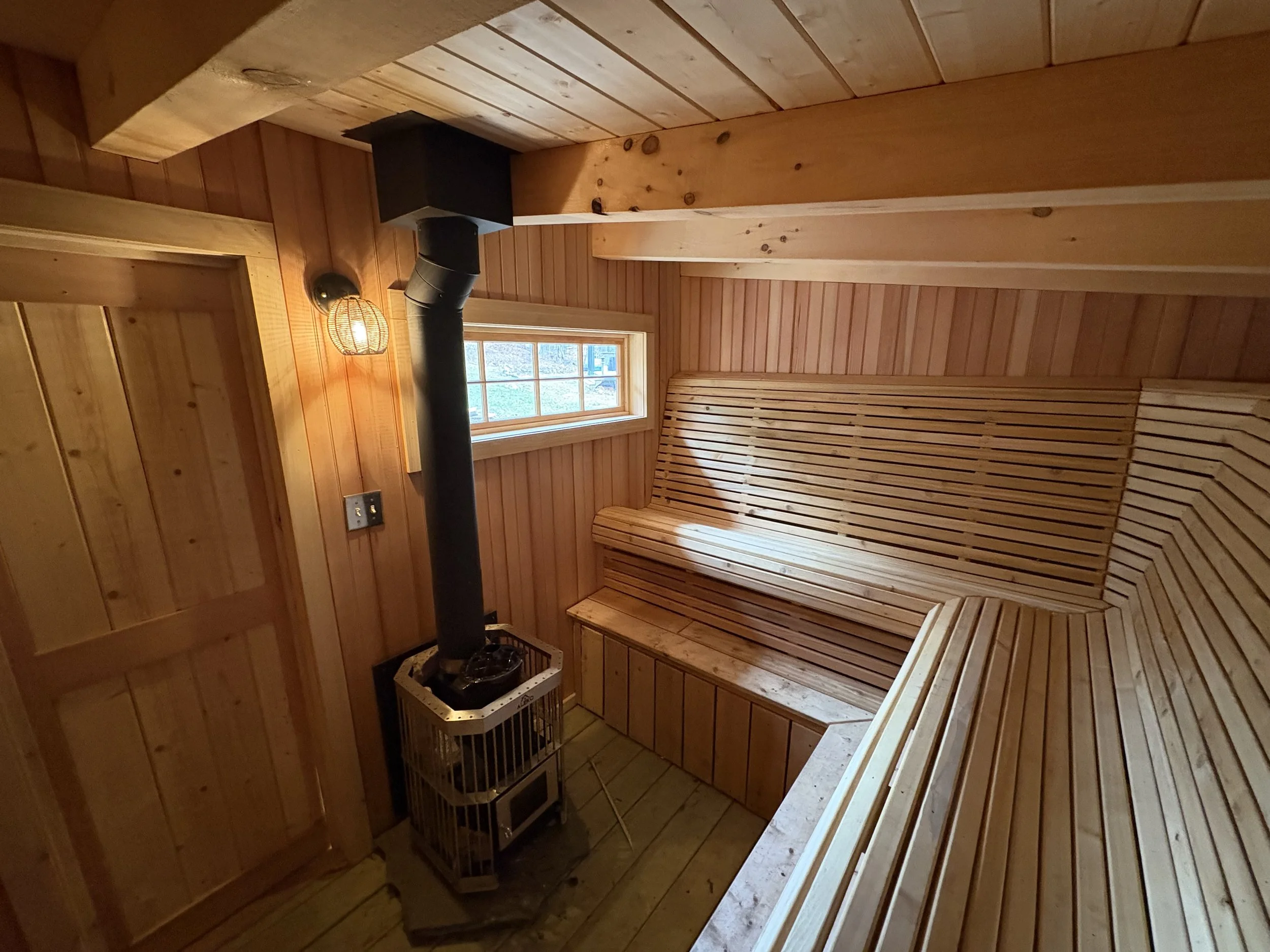 Interior of a wooden sauna room with a bench, a small window, a wall light, and a stove with a chimney pipe. Sauna built by Silas Miller Stove & Chimney