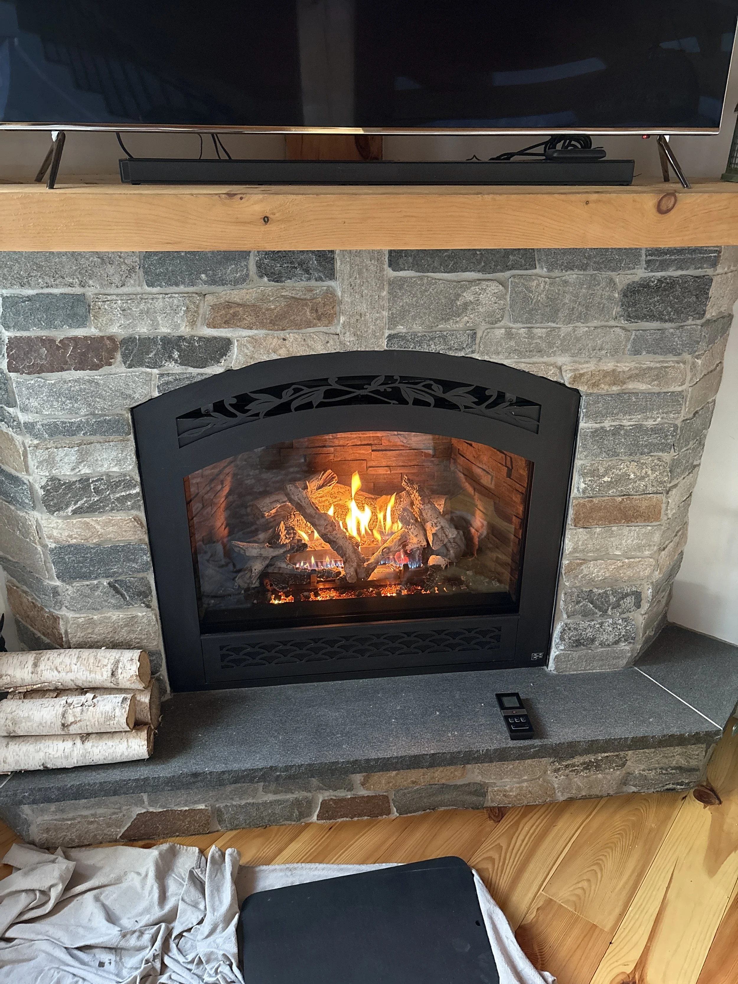 A gas fireplace with a stone surround and a black metal frame with a glass door, burning with logs inside. A remote control is on the hearth, and a few birch logs are stacked to the left of the fireplace. Installed by Silas Miller Stove & Chimney