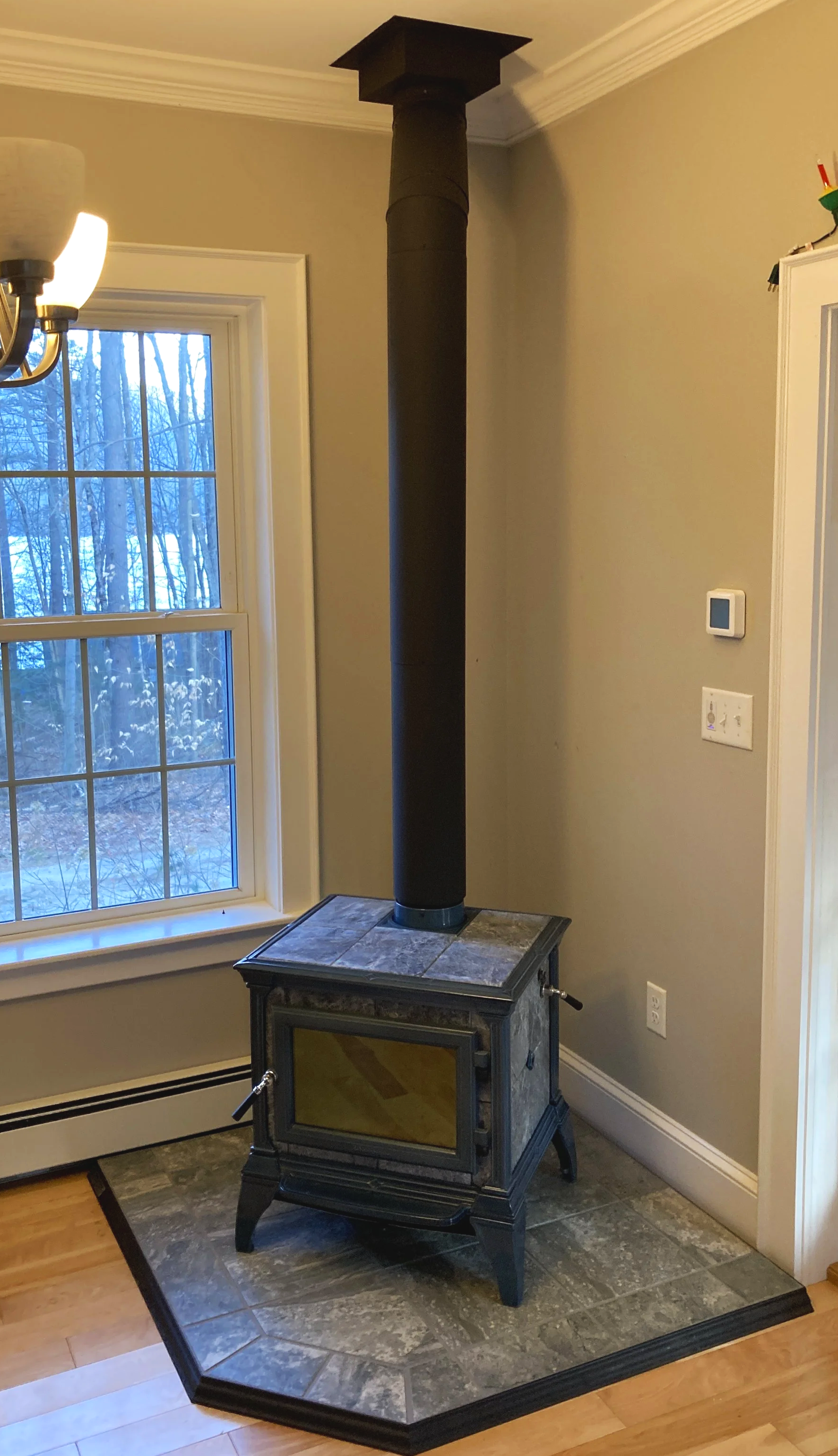 A wood stove with a black chimney pipe in a room with hardwood floors and beige walls, near a window showing a wintery outdoor scene. Silas Miller Stove & Chimney