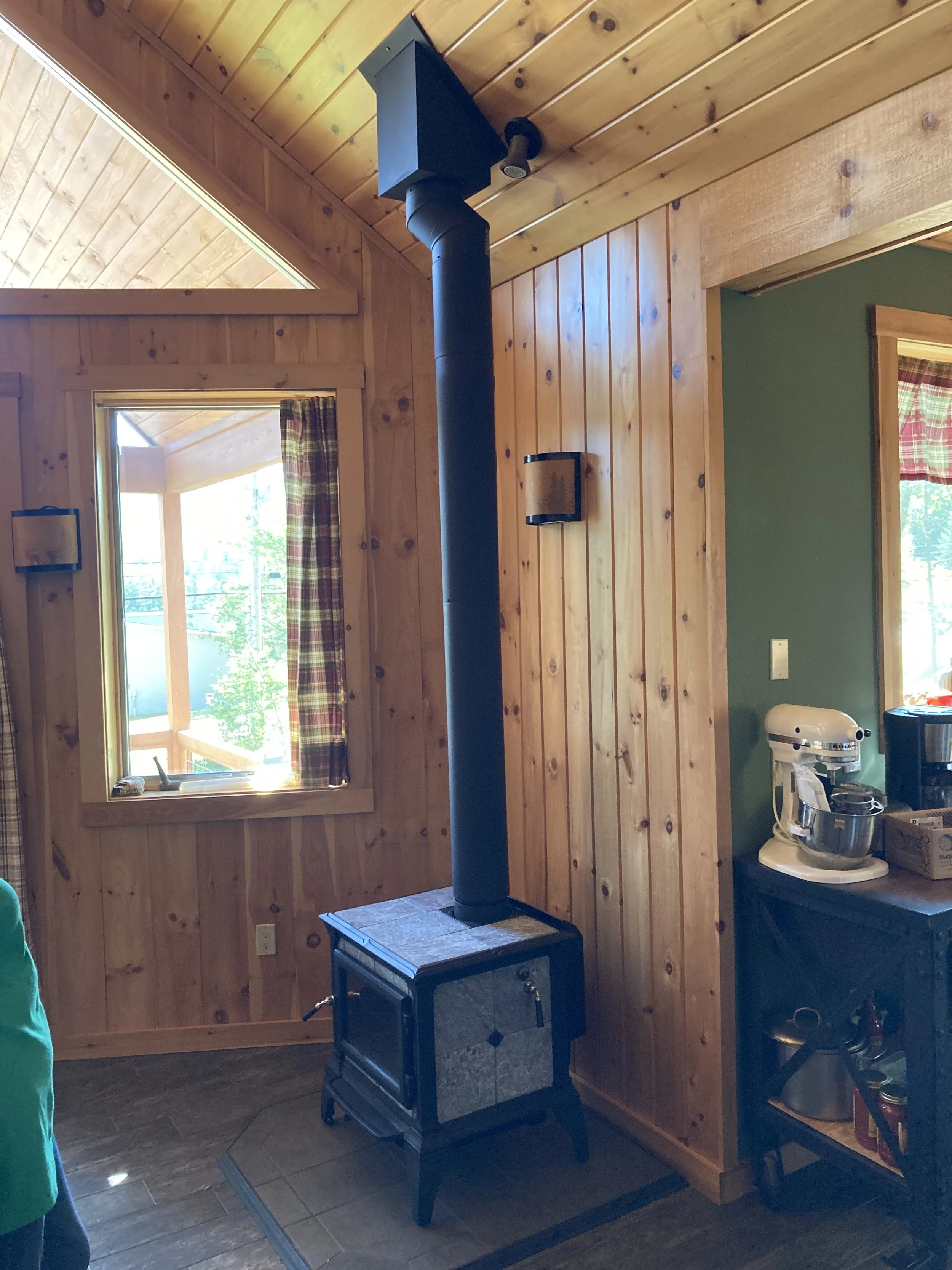 Hearthstone Wood stove with black chimney pipe in a cozy wooden cabin interior with windows and colorful curtains. Silas Miller Stove & Chimney