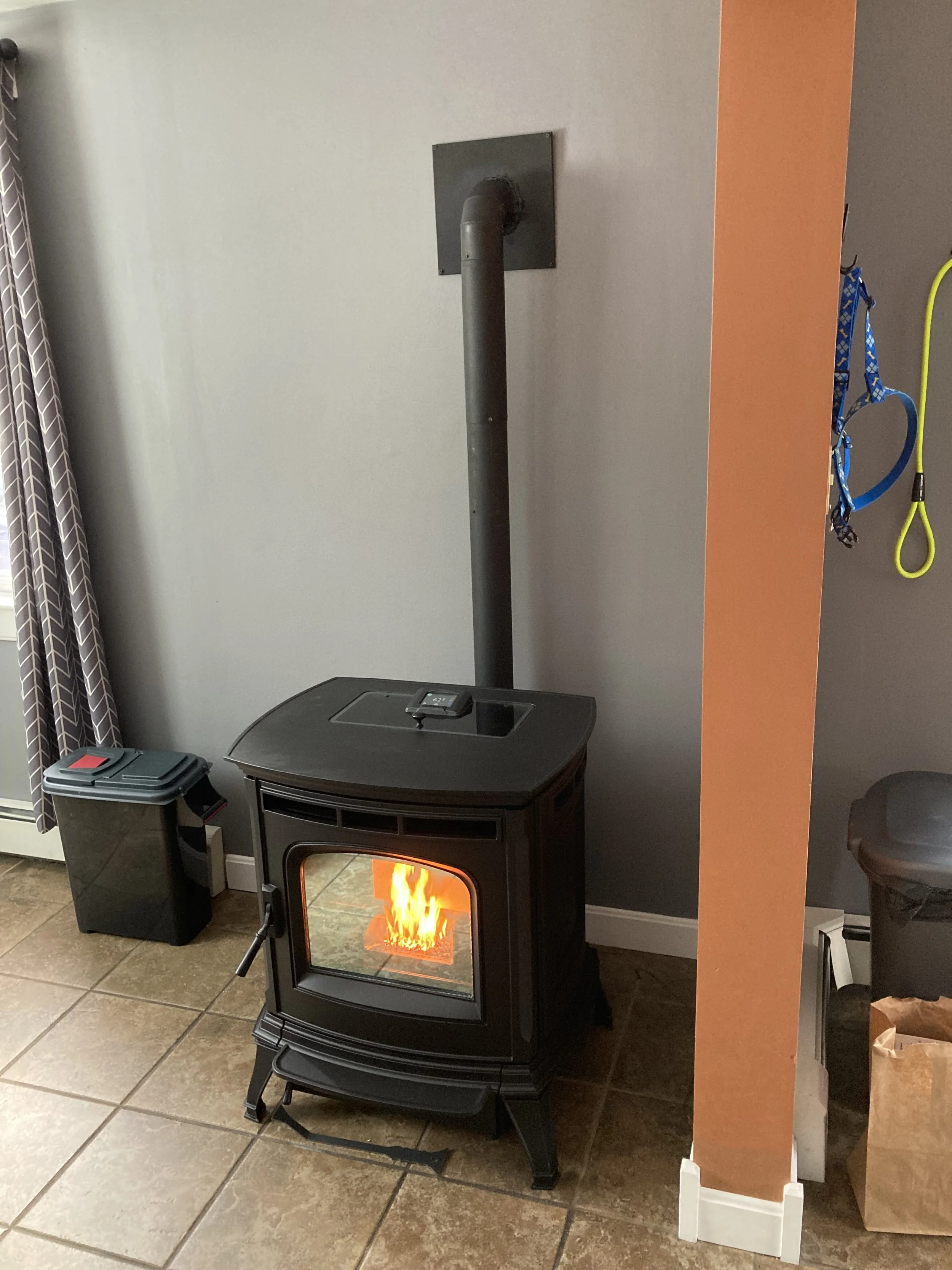 Black harman absolute pellet stove with a fire inside placed on a tiled floor in a room, with a gray wall and a small trash bin nearby. Silas Miller Stove & Chimney