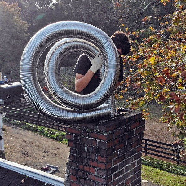 Person standing on a brick chimney holding a large flexible metal liner coil with autumn trees in the background.