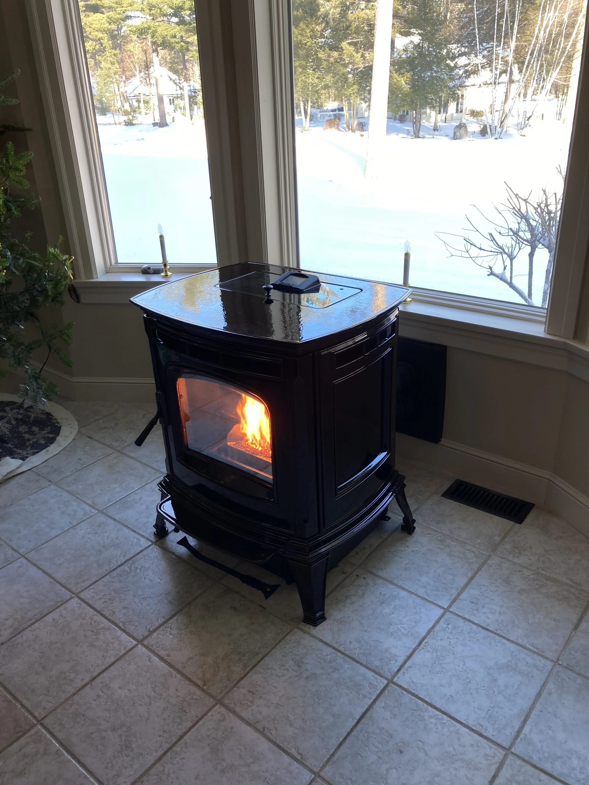 A Harman Absolute pellet stove with an orange fire inside, situated near large windows revealing a snowy outdoor landscape with trees and bushes, and a tiled floor inside. Silas Miller Stove & Chimney