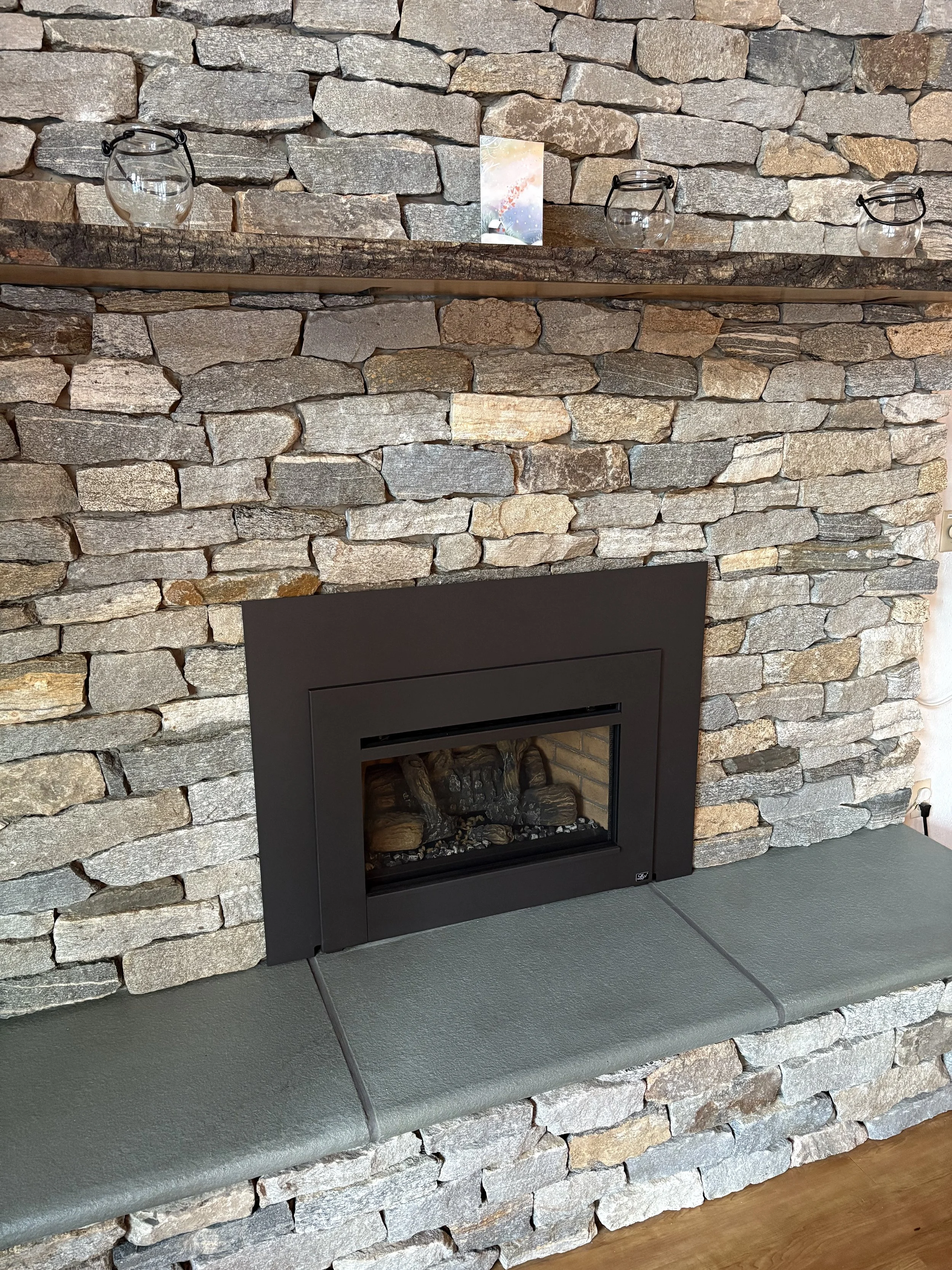 A stone fireplace with a black rectangular gas insert, a stone hearth, a wooden mantel, and three glass candle holders on the mantel. Installed by Silas Miller Stove & Chimney