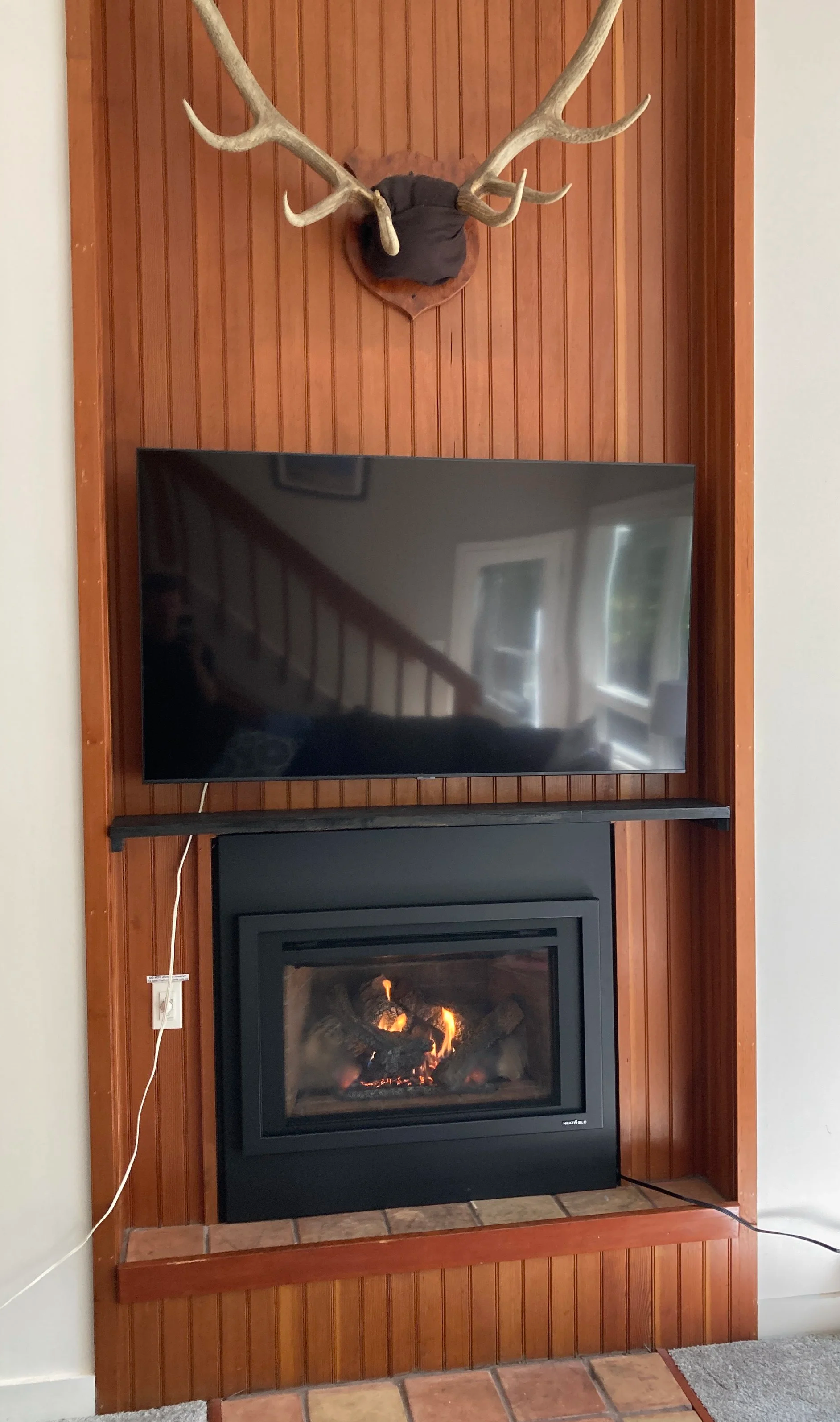 Living room gas fireplace with a mounted deer head above, and television, with a fireplace below showing lit logs. Silas Miller Stove & Chimney