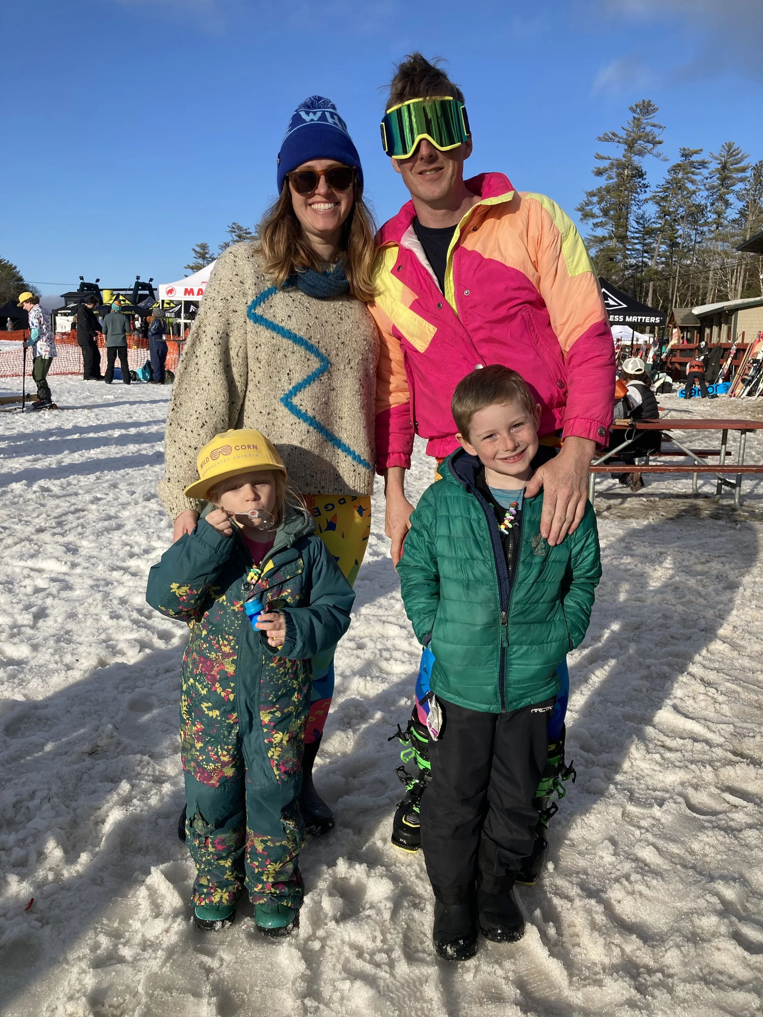 Silas Miller and family of four with two kids and two adults posing on a snowy day at a ski resort or snow park. The woman is wearing a beige sweater, sunglasses, and a knit hat, while the man is wearing ski goggles and a colorful ski jacket.