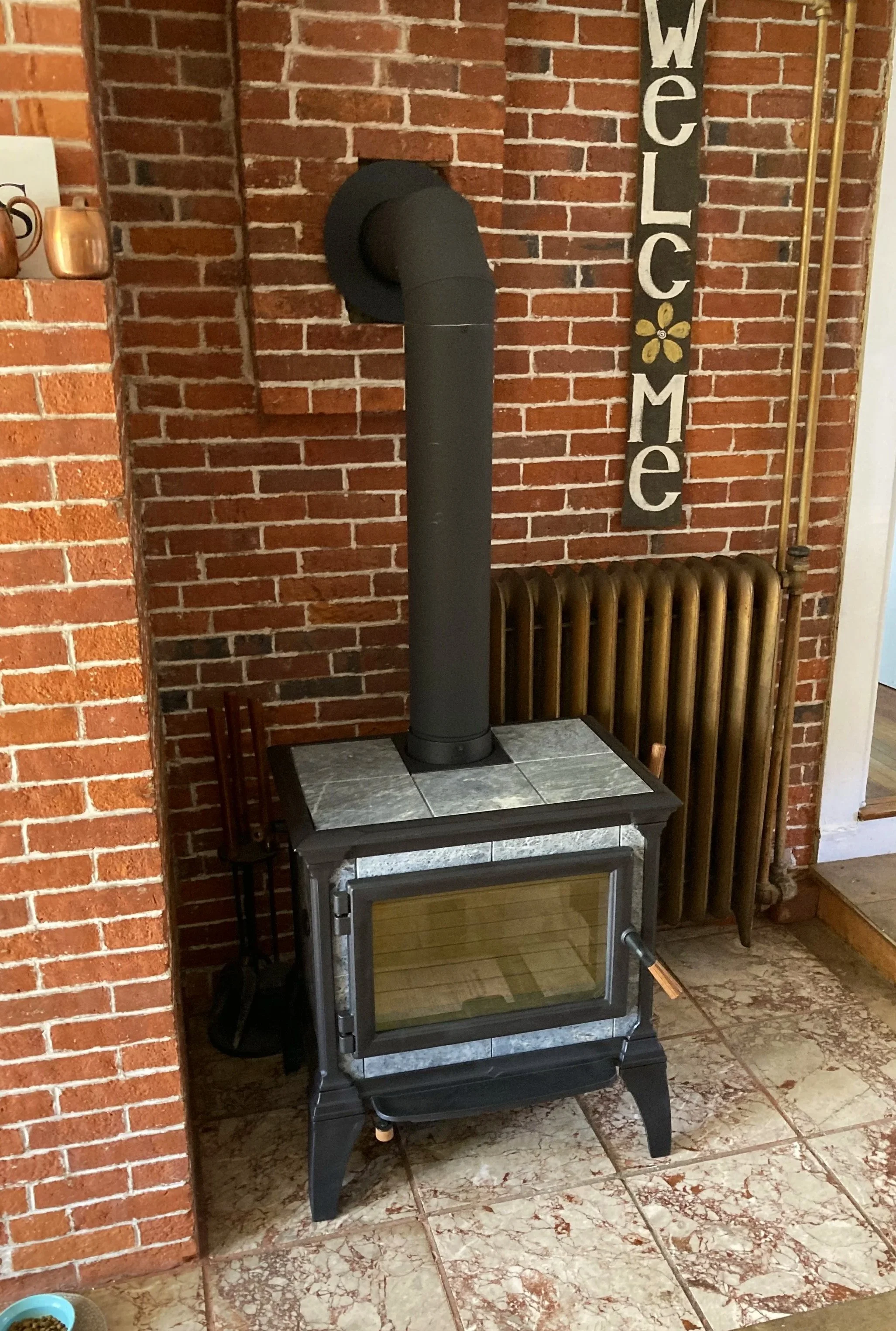 A Hearthstone soapstone wood stove, positioned against a red brick wall in a cozy interior space. To the right, a "Welcome" sign hangs on the wall, and a vintage radiator is visible behind the stove. Silas Miller Stove & Chimney