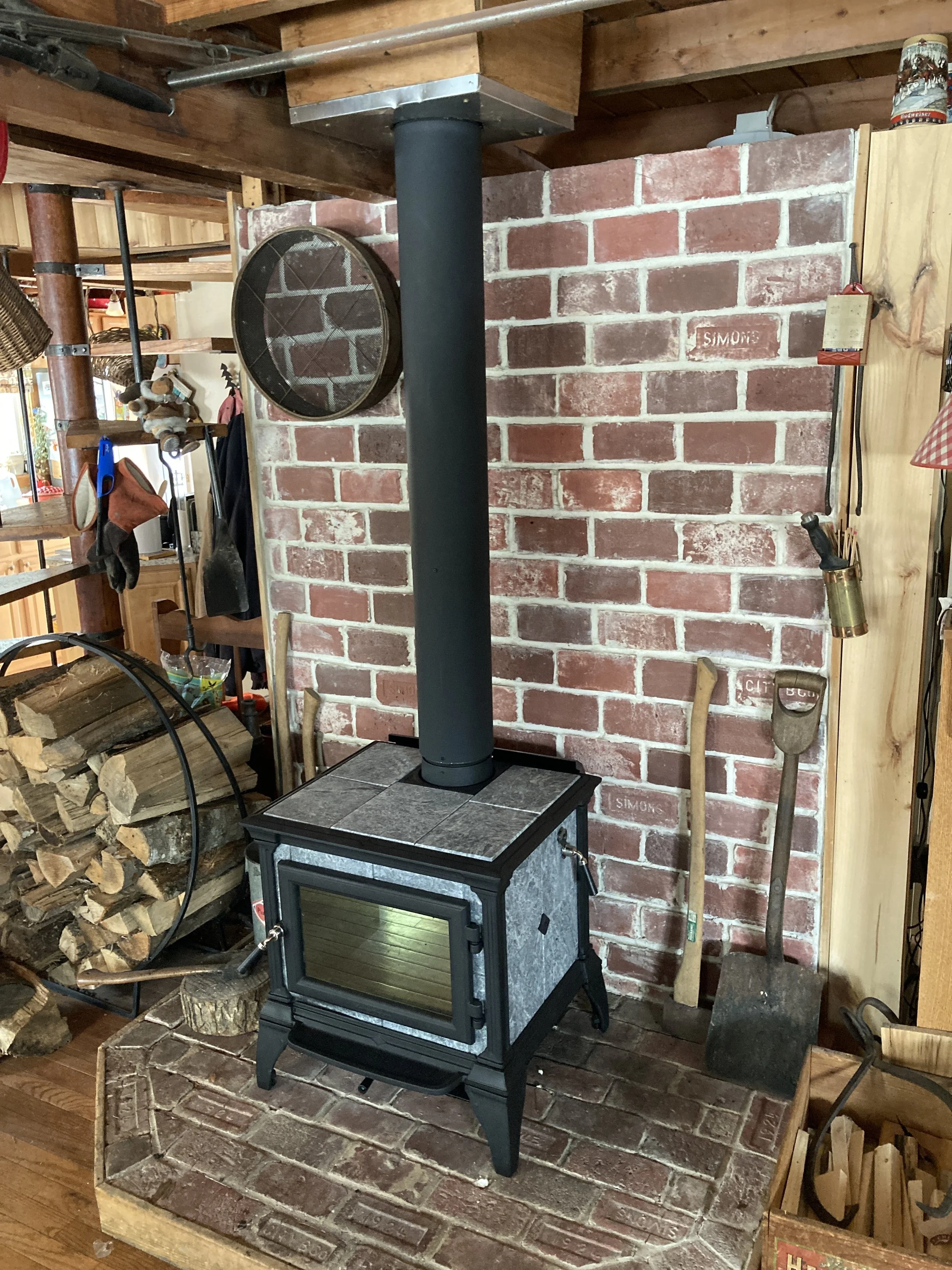 A wood-burning stove with a black chimney pipe against a red brick wall, with winter tools like an axe and shovel nearby, in a cozy rustic interior. Installed by Silas Miller Stove & Chimney