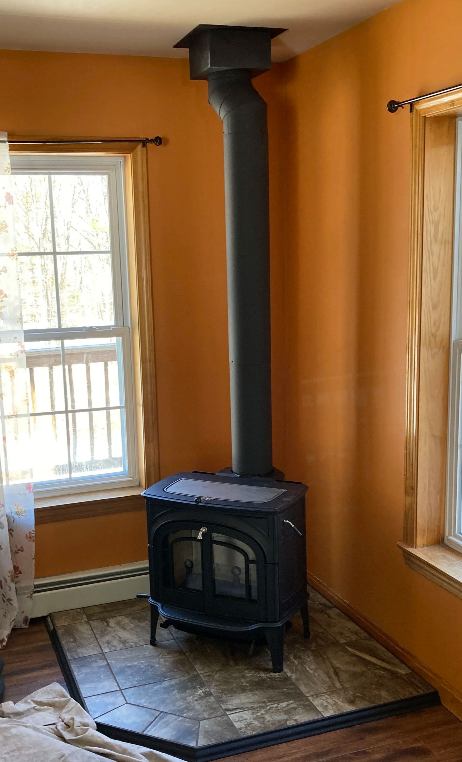 A black wood-burning stove in a corner of a room with orange walls and wooden window frames, sitting on a tiled hearth, with a stovepipe chimney extending upward to the ceiling. Silas Miller Stove & Chimney