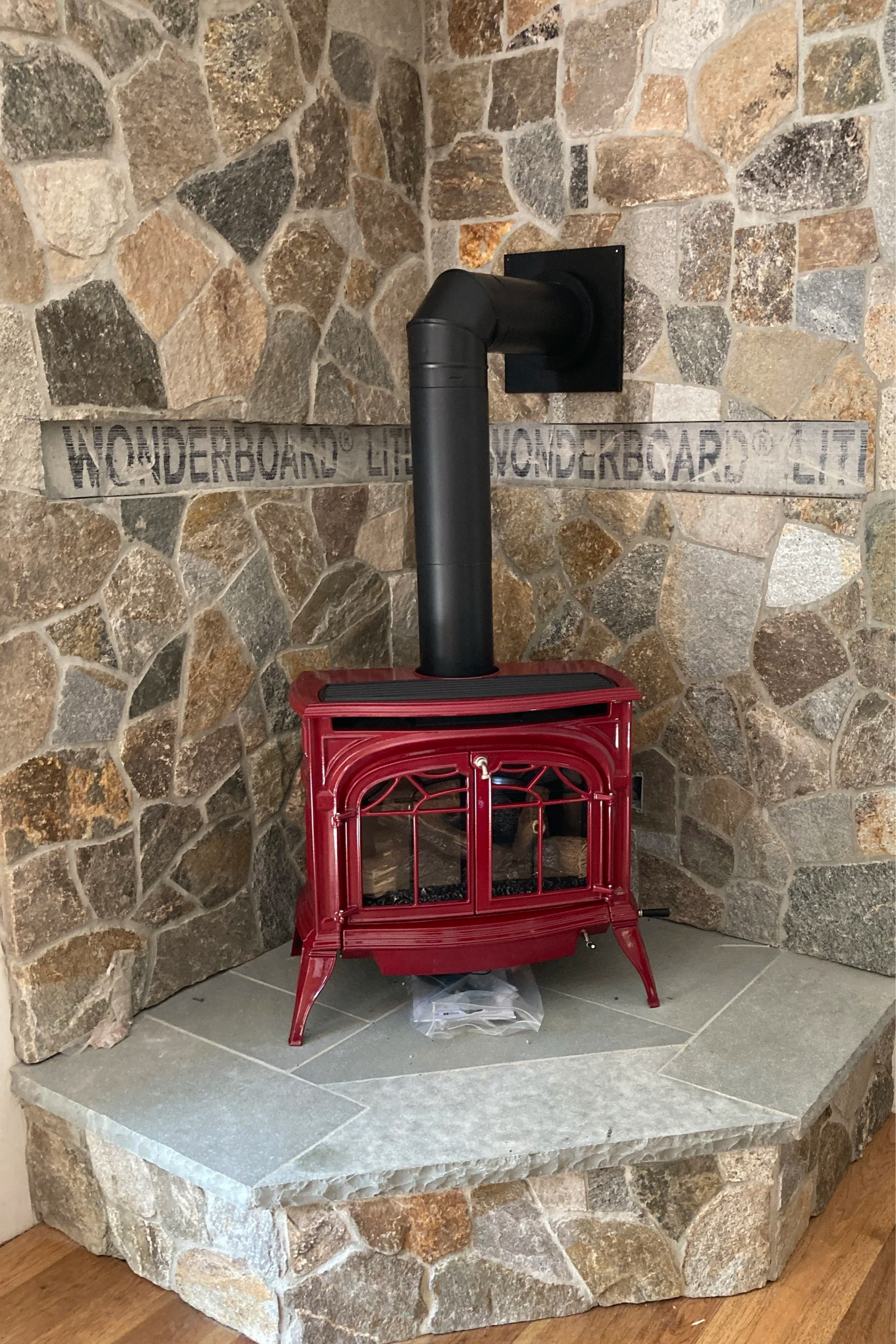 Red cast iron gas stove with black chimney pipe in front of a stone fireplace wall. Silas Miller Stove & Chimney