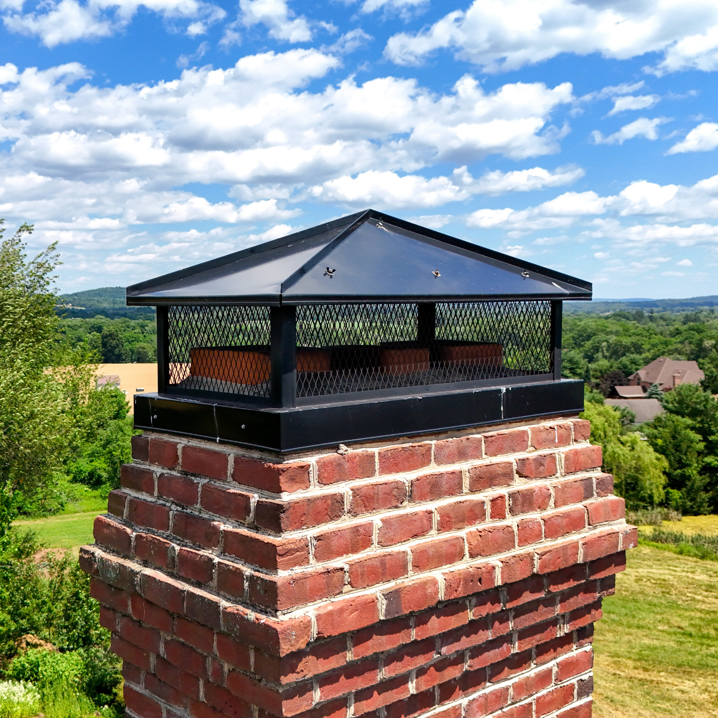 Close-up of a brick chimney with a black metal chimney cap and spark guard, set against a bright blue sky with scattered clouds and green landscape in the background.