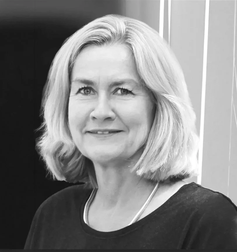 Schwarz-Weiß-Fotografie einer lächelnden Frau mit kurzer, blonder Haarfrisur, die eine dunkle Oberbekleidung trägt.