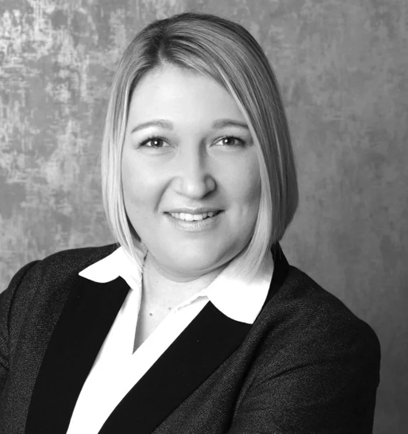 Schwarz-weiß Porträt einer lachenden Frau mit kurzen Haaren, im Business-Look, vor einem gemusterten Hintergrund.