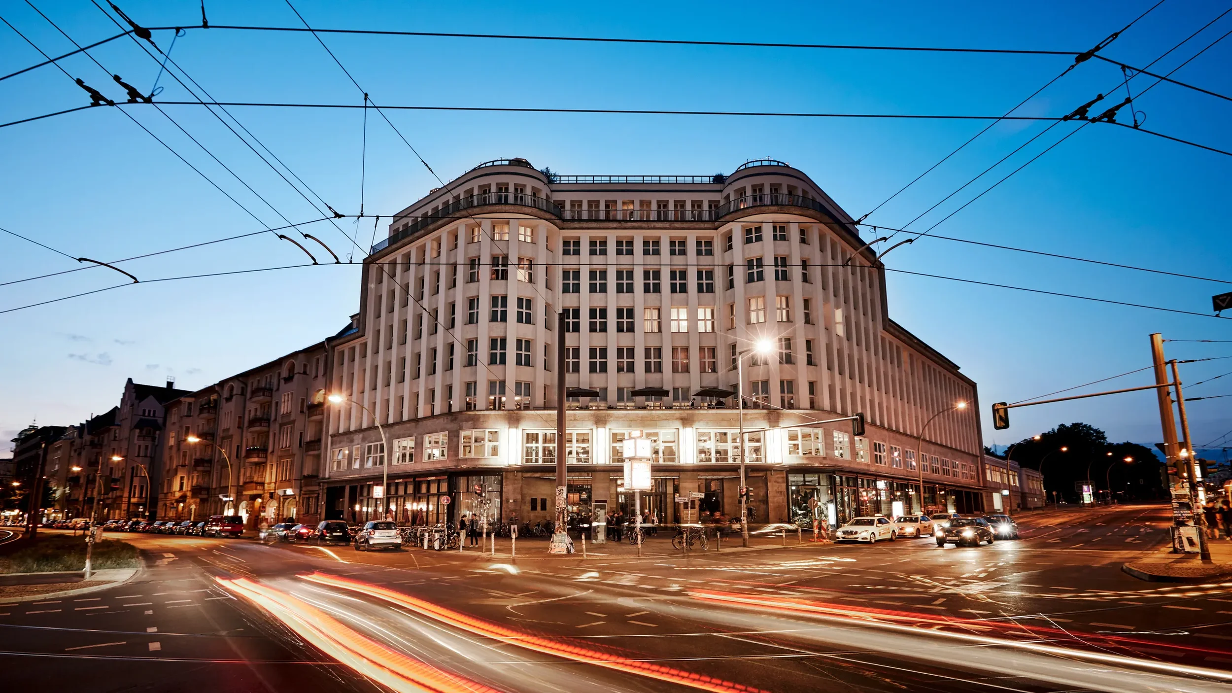 Ein mehrstöckiges Gebäude an einer belebten Straßenecke in der Stadt bei Dämmerung, mit beleuchteten Fenstern und Verkehrsampeln.