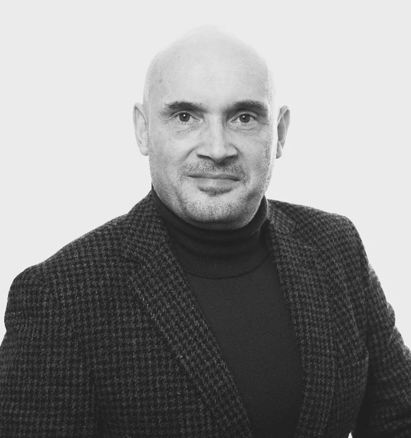 Schwarz-Weiß-Porträt eines Mannes mit Glatze, dunklem Turtleneck und kariertem Blazer vor hellem Hintergrund.