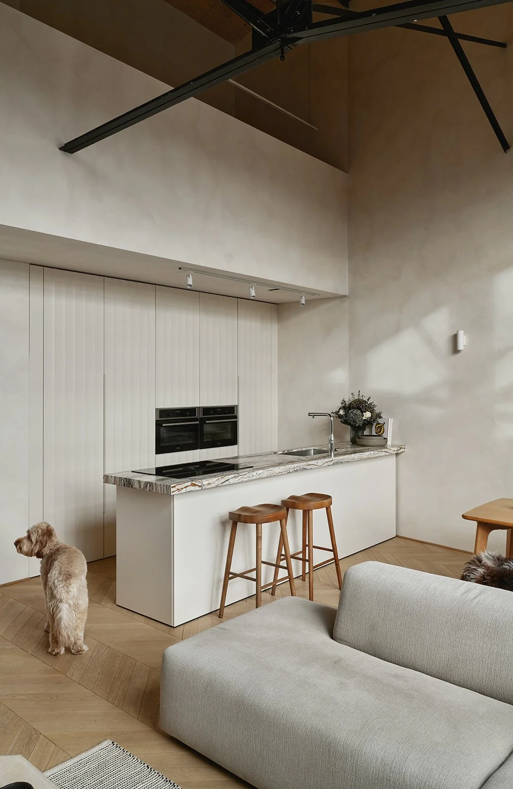 A modern kitchen with a marble countertop, two wooden stools, and a floral centerpiece. There's a beige couch and a dog visible in the living area.