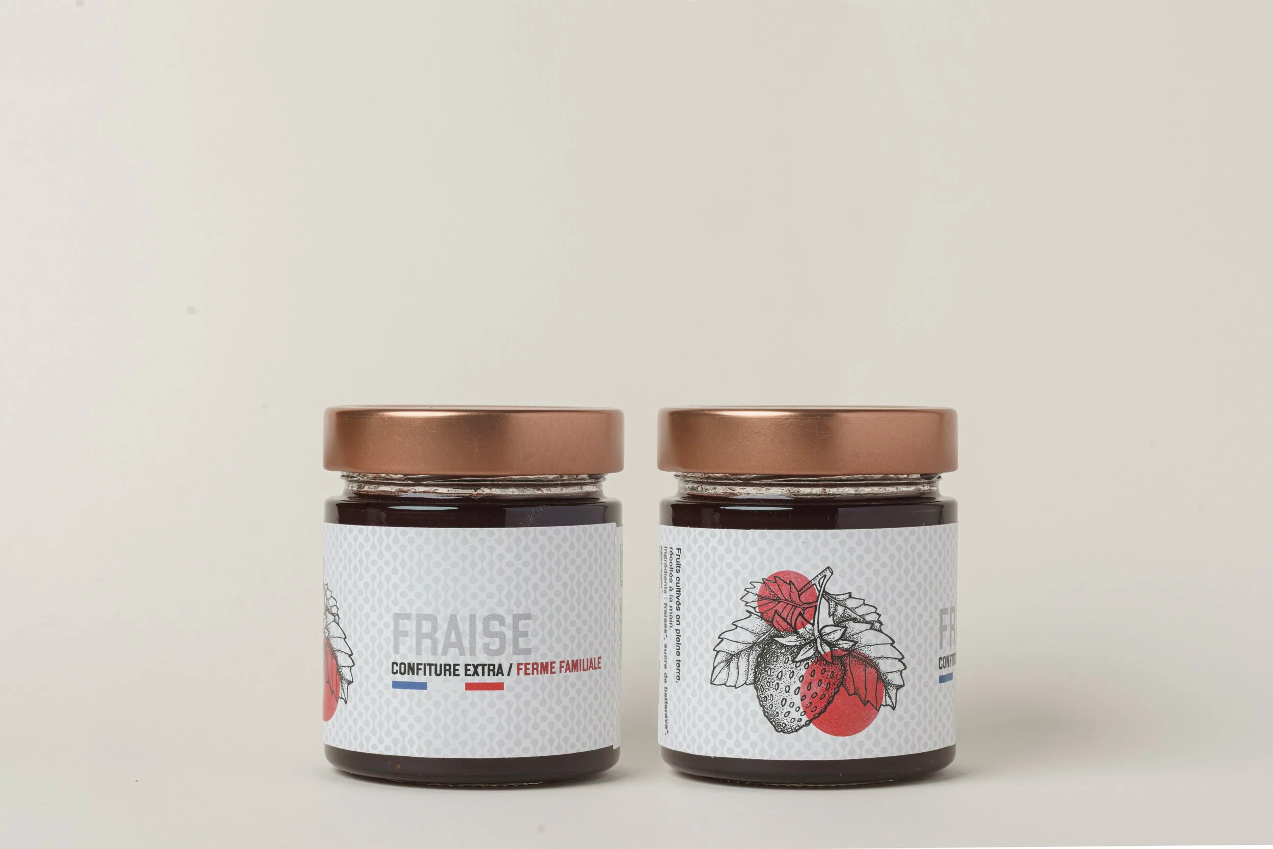Two jars of strawberry jam with white labels and copper-colored lids are placed side by side against a plain off-white background.