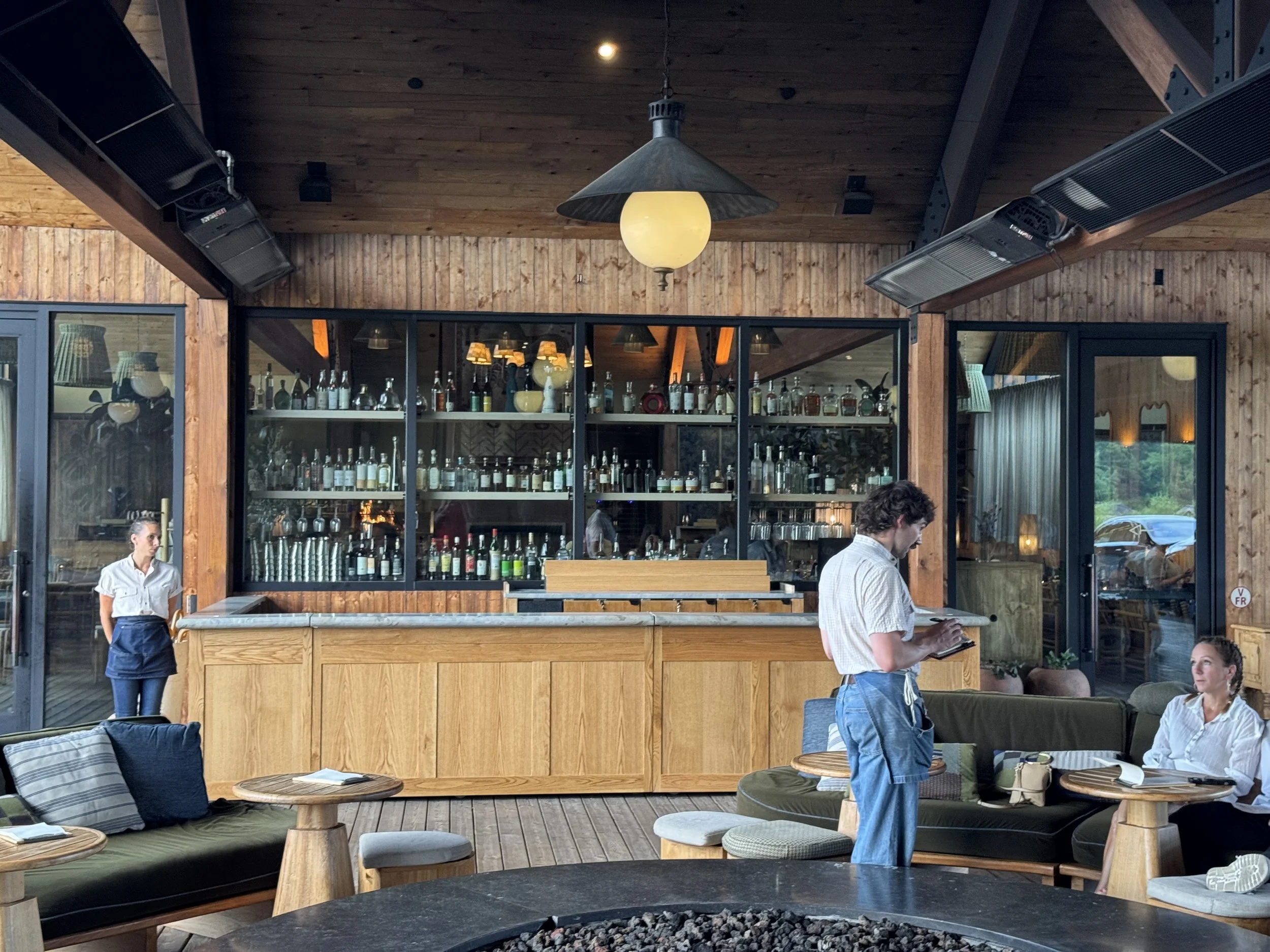 Interior of a rustic restaurant or cafe with wooden walls and ceiling, large glass windows, and a bar stocked with bottles. There are several people including a woman in a white shirt and a man reading a menu. The space has cozy seating with cushions