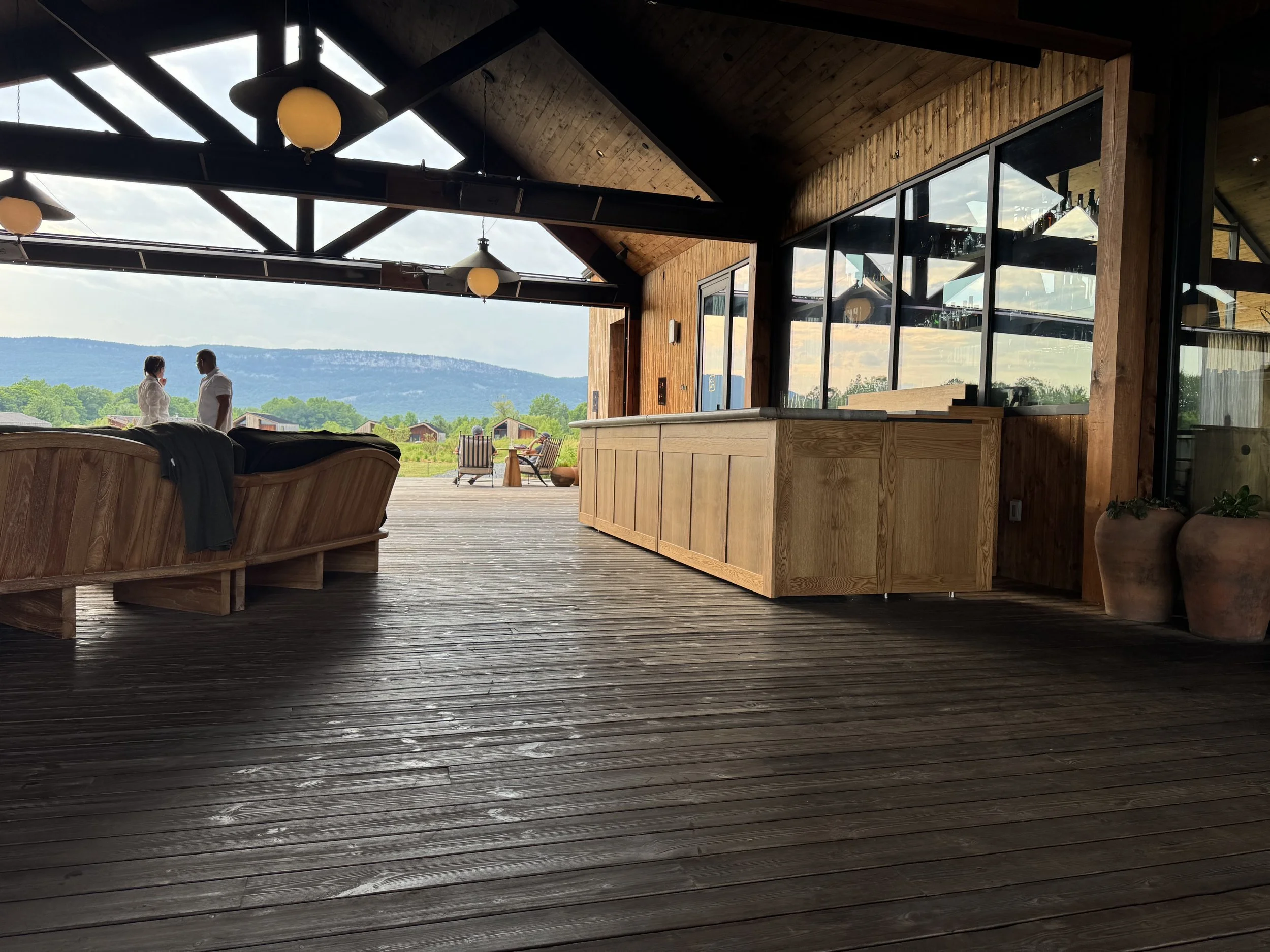 Interior view of a modern restaurant or lounge with wooden walls and ceiling, large glass windows overlooking a scenic landscape, with two people talking near the patio, outdoor chairs, and ceiling lights.