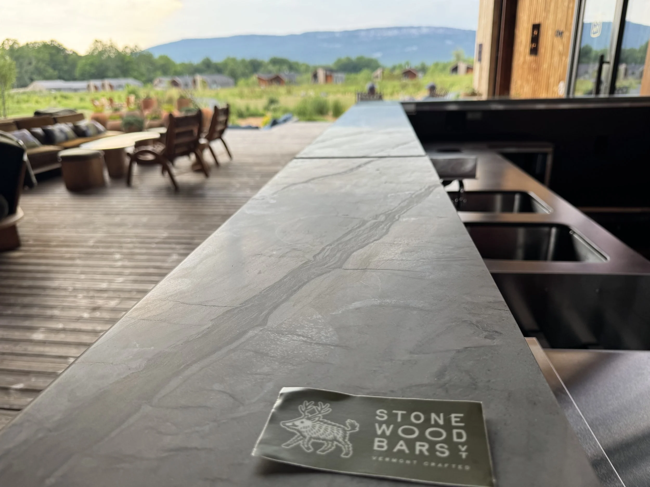 A view from inside a kitchen showing a marble countertop with a sink, overlooking an outdoor patio with chairs and tables, and a rural landscape with green fields, small buildings, and mountains in the distance.