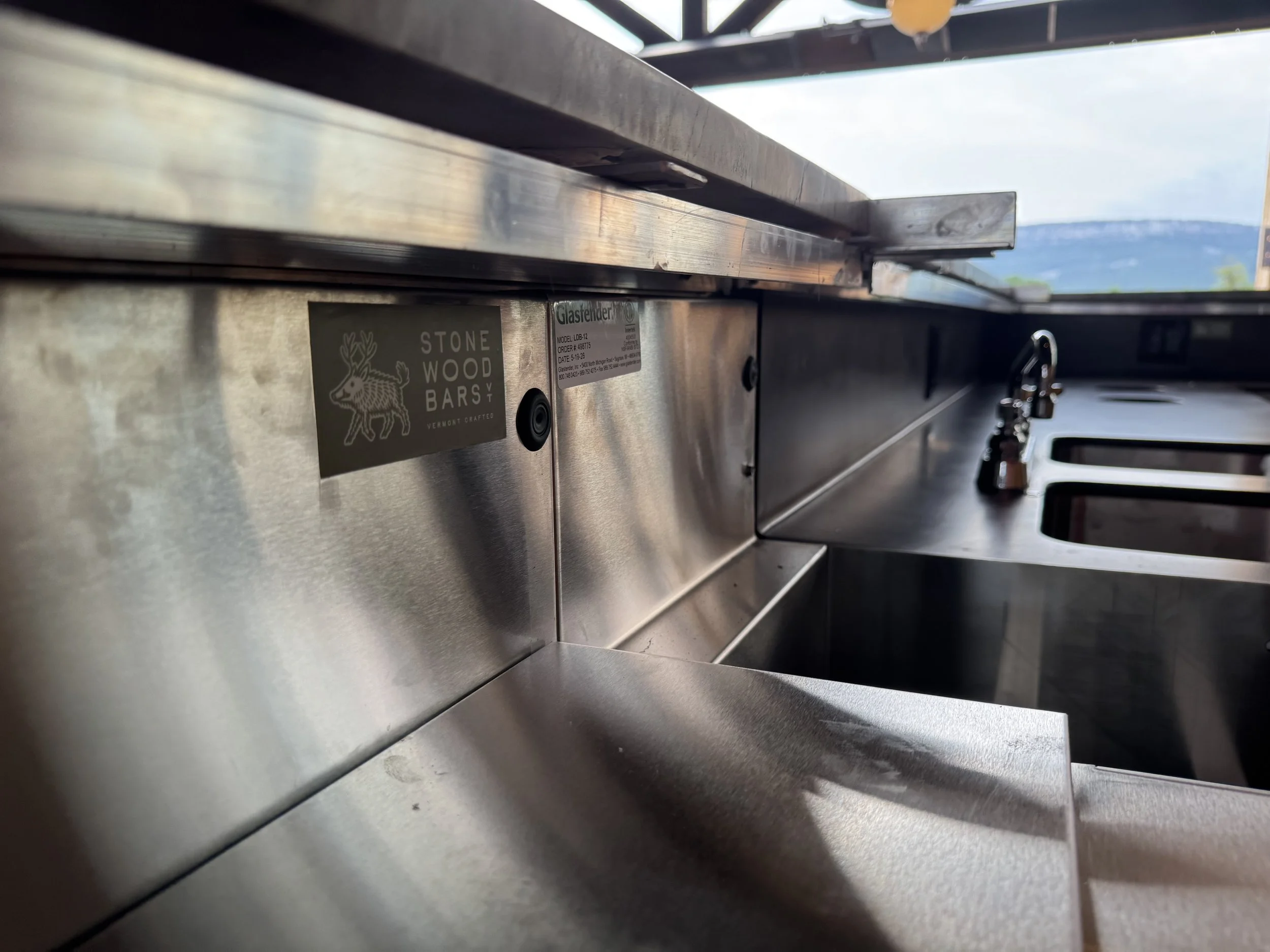 Close-up of a stainless steel kitchen counter with a sticker that says "Stone Wood Bar" and a drawing of a reindeer, with a window showing a mountain landscape in the background.