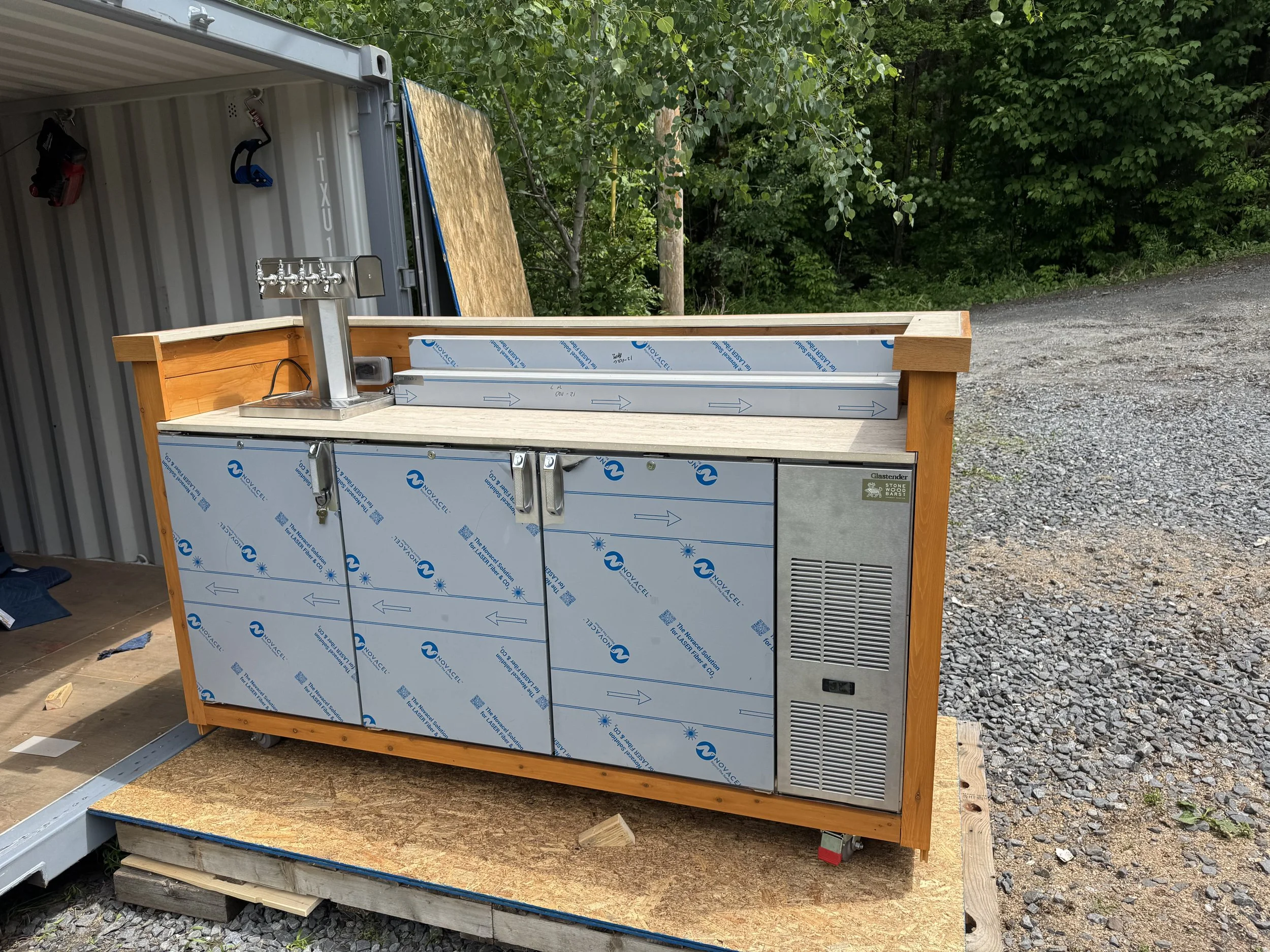 A construction site shows a rectangular outdoor bar made of wood and metal, with seven taps on one side and a grid for refrigeration on another. The bar is partially covered with protective film, and there's a wood platform beneath it on gravel groun