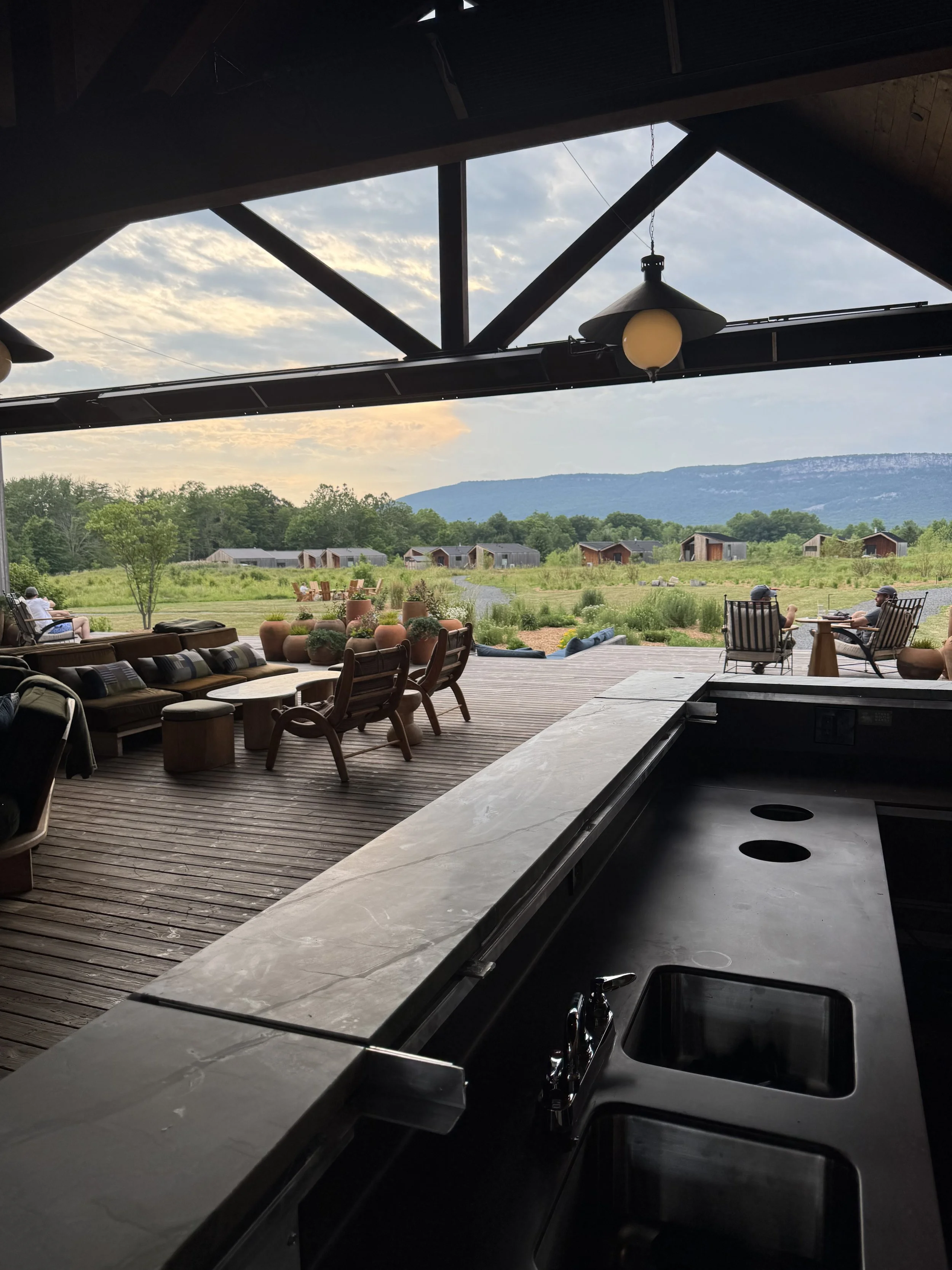 A view from inside a kitchen looking out onto a deck and grassy field with small wooden buildings and mountains in the background. The kitchen features a black countertop with a sink and stovetop, and a window opening to the outdoor seating area with