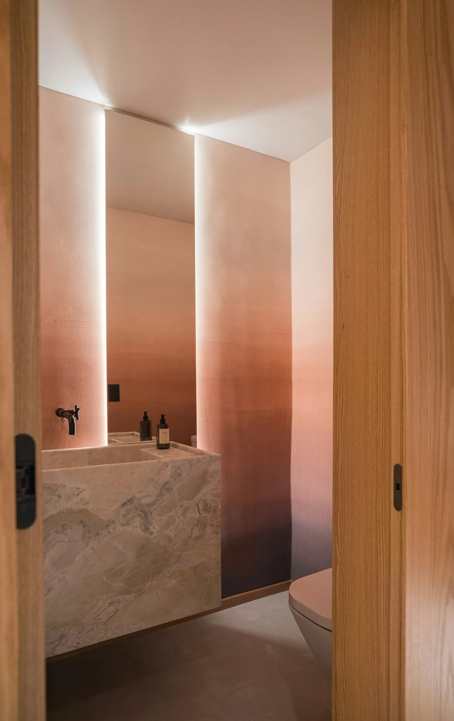 A minimalist bathroom with a pink-toned wall, a marble vanity with a dark faucet, and a white toilet partially visible, viewed through a wooden sliding door.