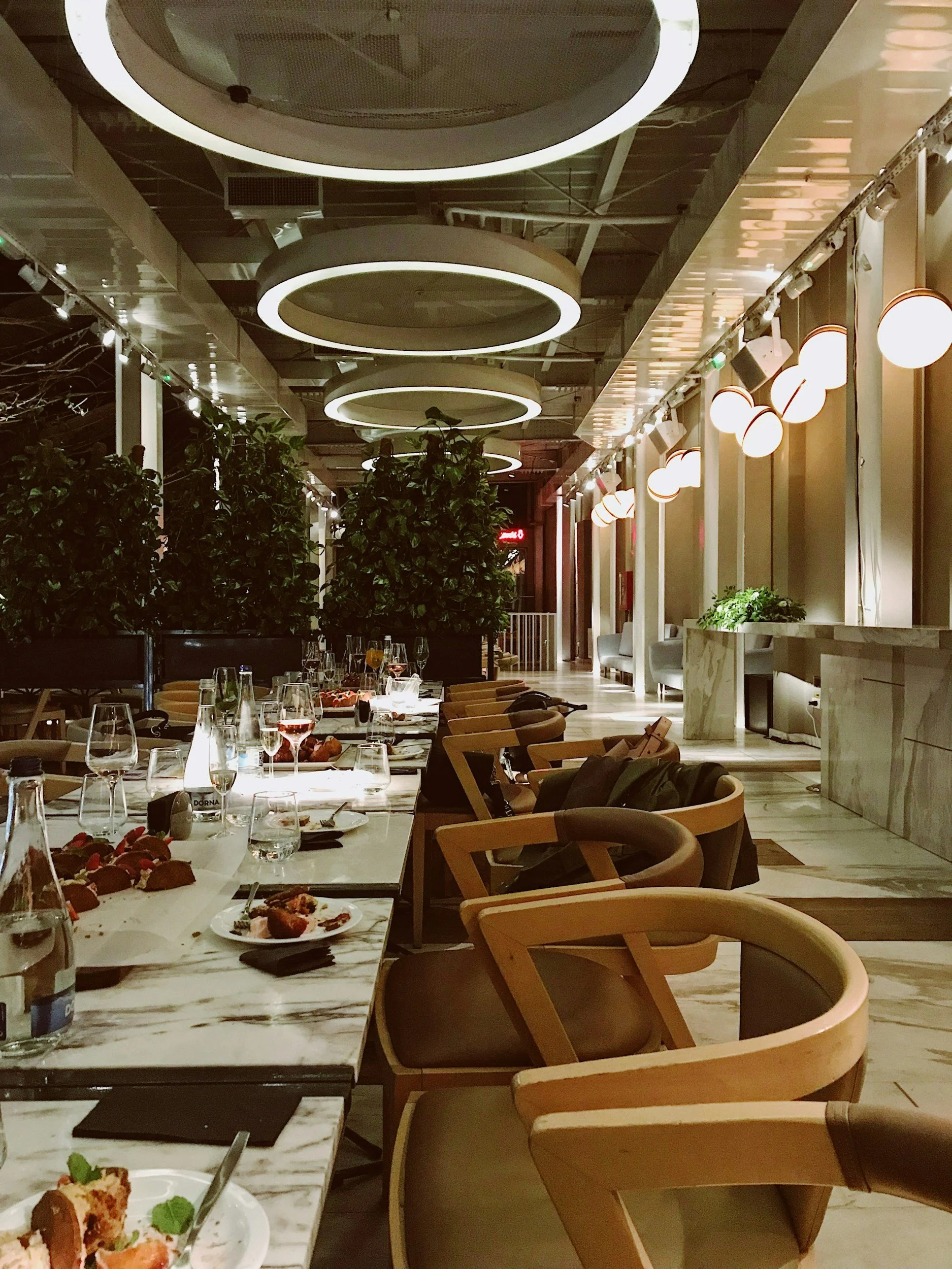 Interior of a restaurant with a dining table set with plates, glasses, and food. Stylish lighting fixtures hang from the ceiling, and there are chairs around the table. The background has some green plants and a long corridor.