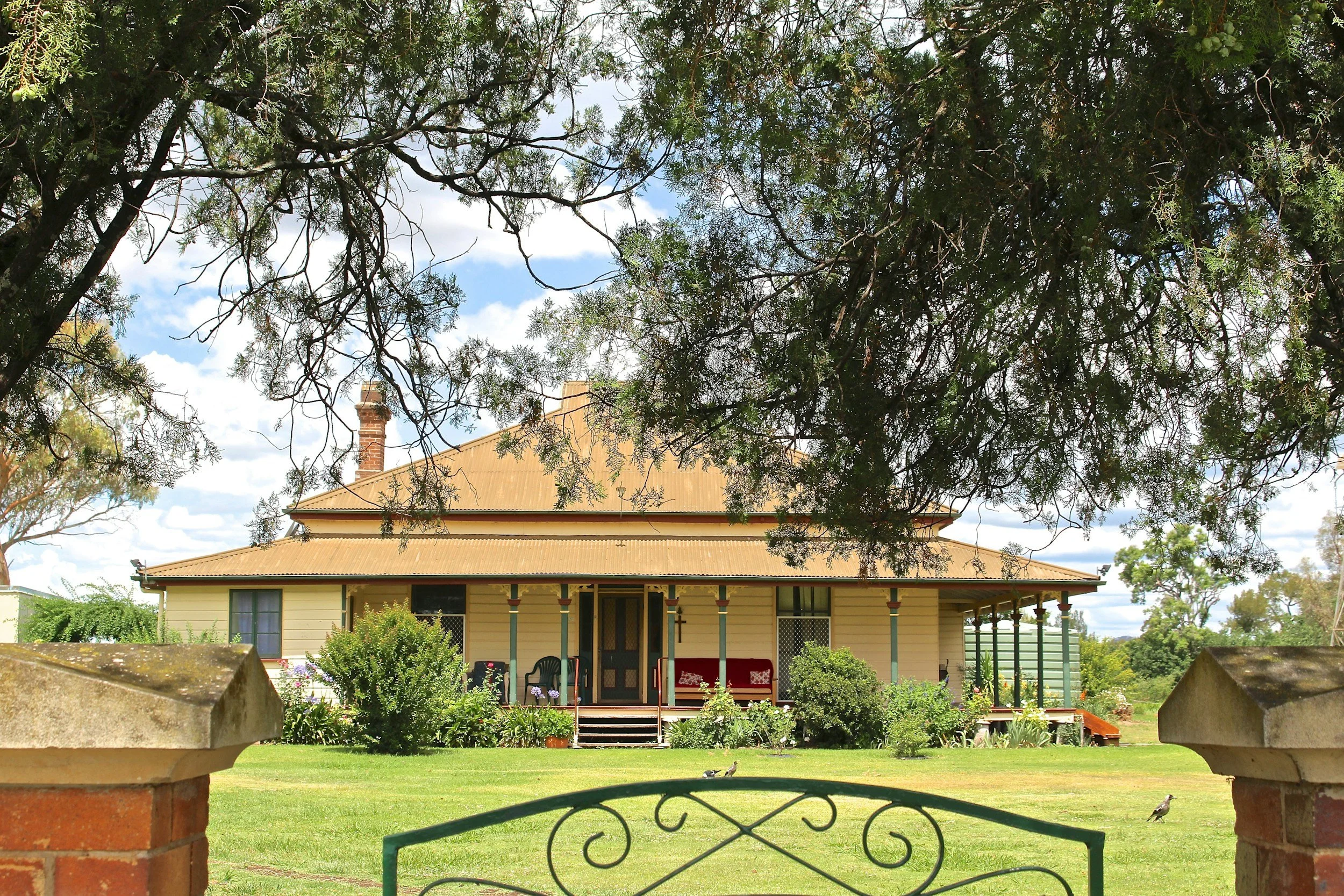 A large, historic, single-story house with a wraparound porch, painted in a light yellow color with green trim. The house has a metal roof and is surrounded by lush, well-maintained garden and trees. A green metal gate is visible in the foreground, with brick pillars on either side, and two small birds on the grass near the gate. The sky is partly cloudy with patches of blue.