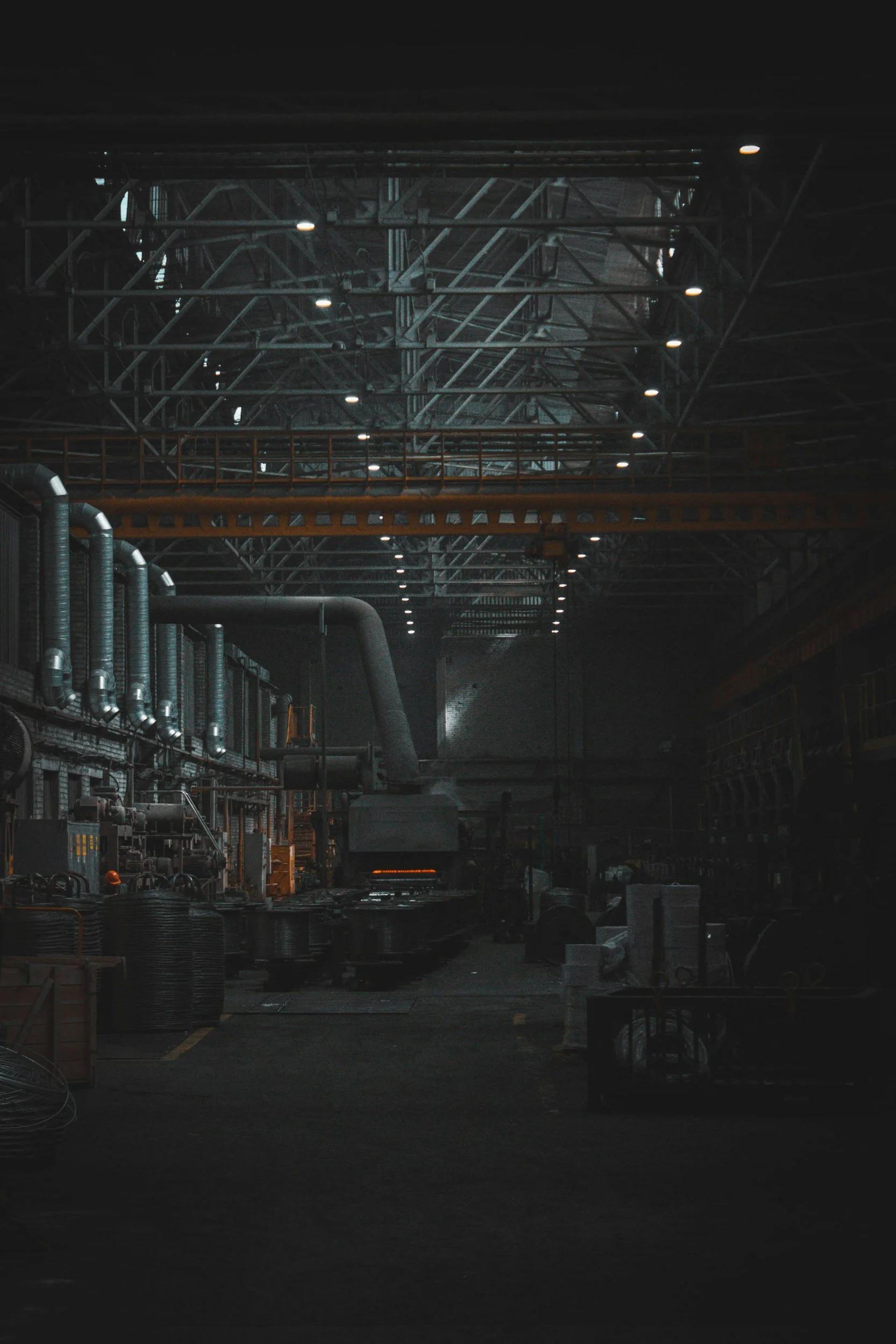 Inside an industrial factory with heavy machinery, pipes, and overhead lighting in a large dark warehouse.