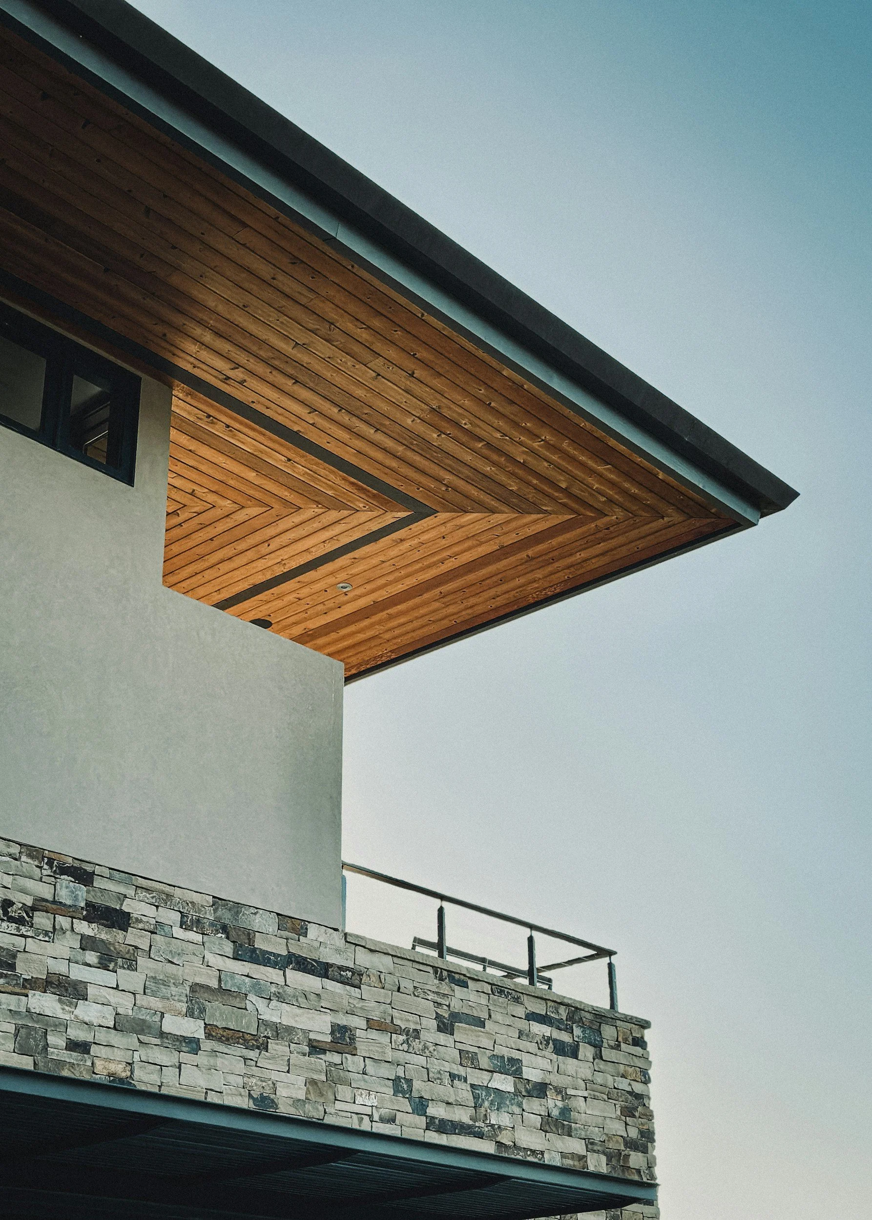 Close-up of a modern house's upper corner with a stone wall, white wall, and wooden ceiling under the roof, against a clear sky.