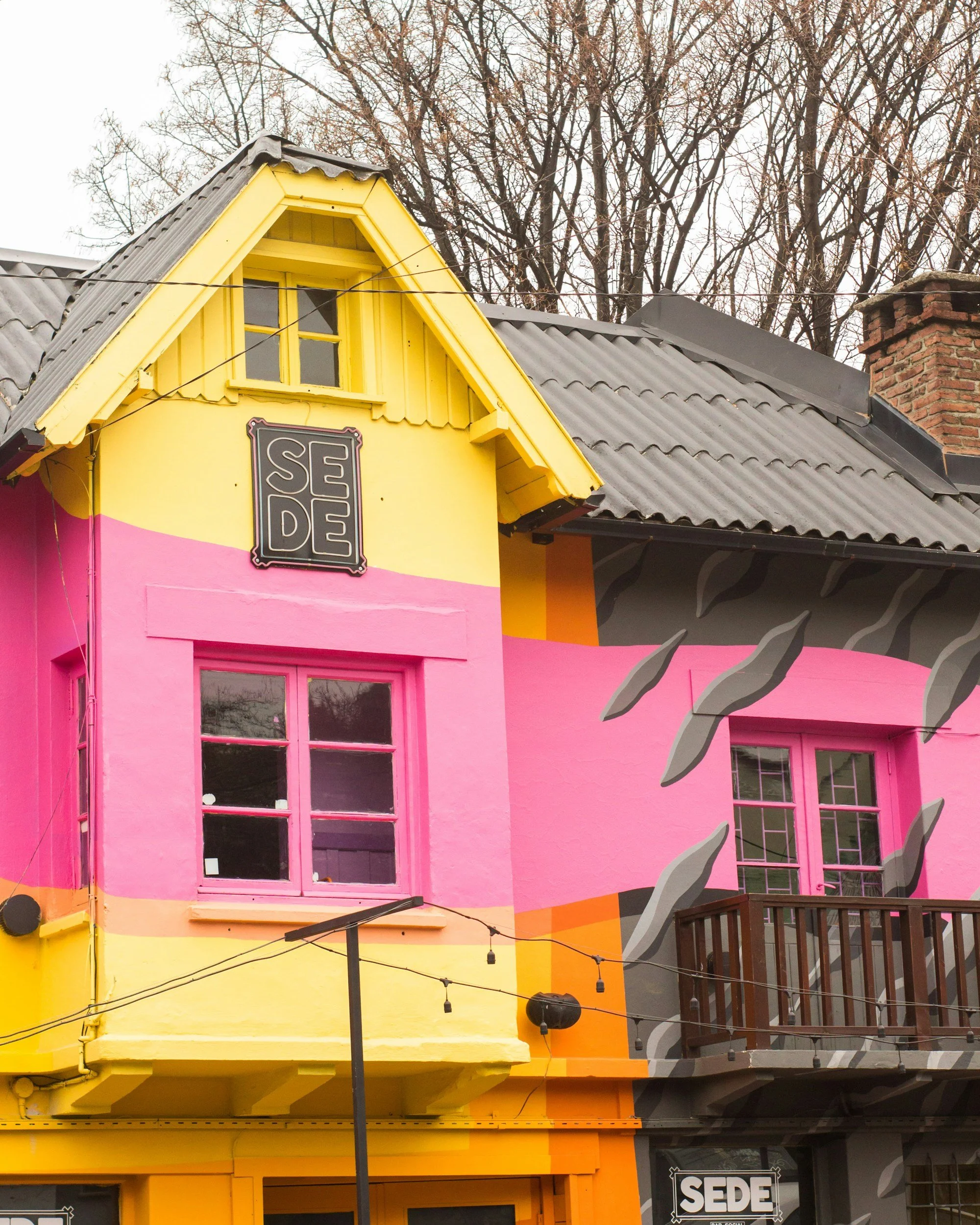 Colorful building with pink and yellow walls, two windows with pink frames, a yellow gable roof, and a sign labeled SEDE. There are leafless trees in the background and string lights hanging in front.