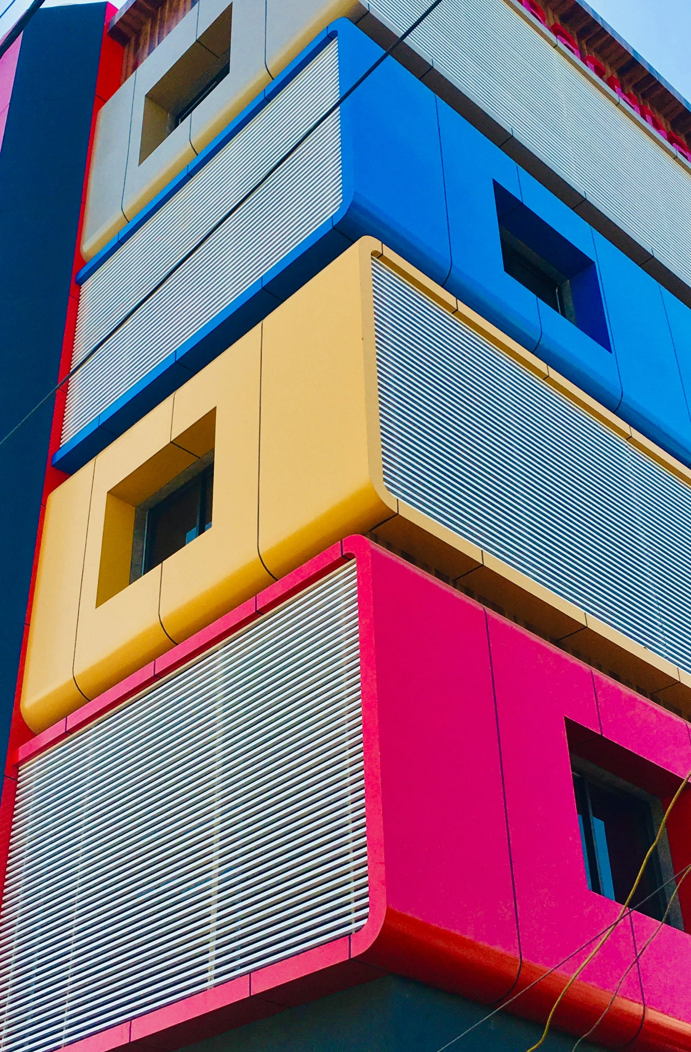 Colorful modern building with yellow, blue, and pink sections, featuring square windows and metallic horizontal louvers.