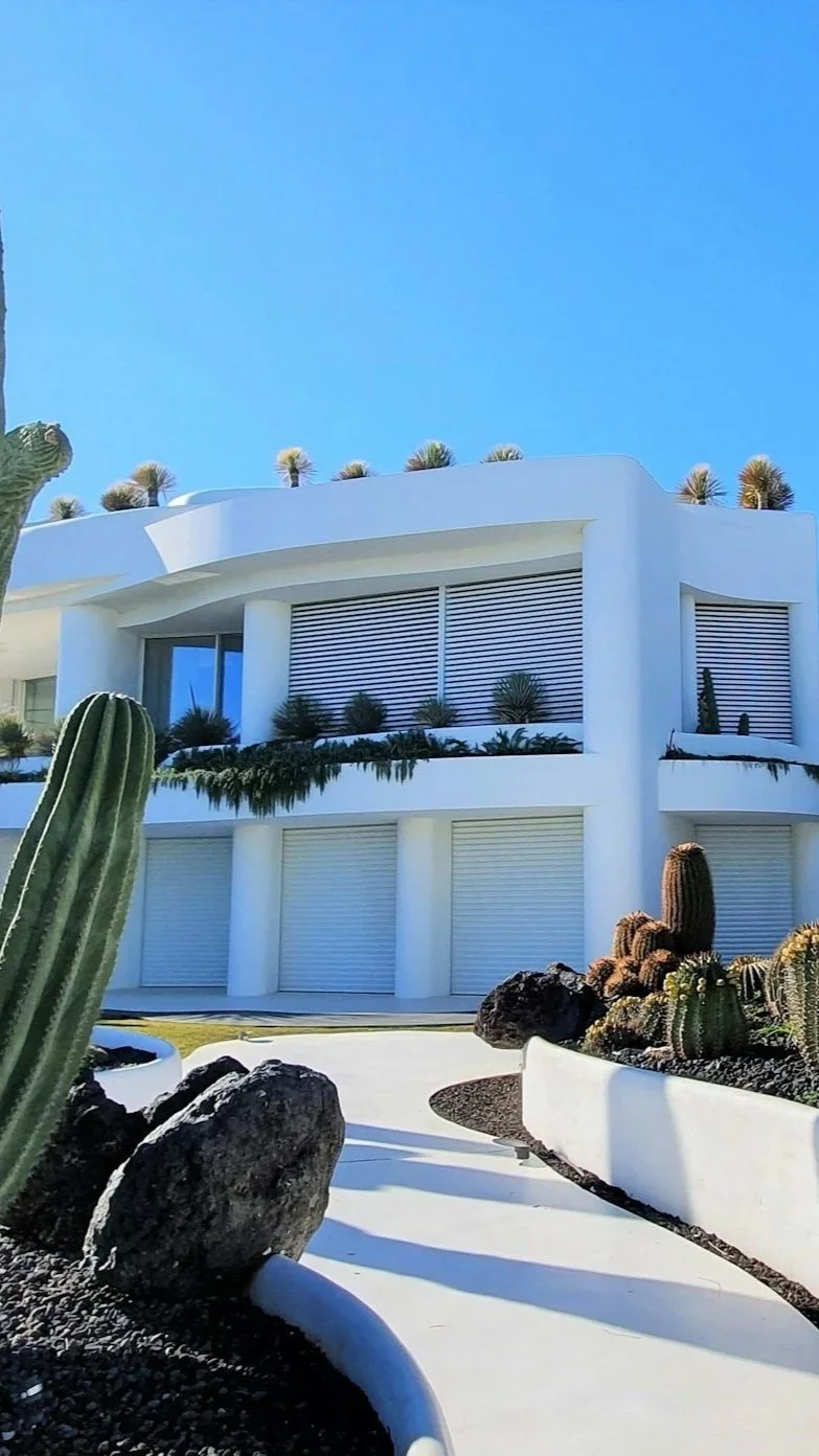 Modern white house with large windows, horizontal blinds, and balconies, surrounded by desert plants and cacti, under a clear blue sky.