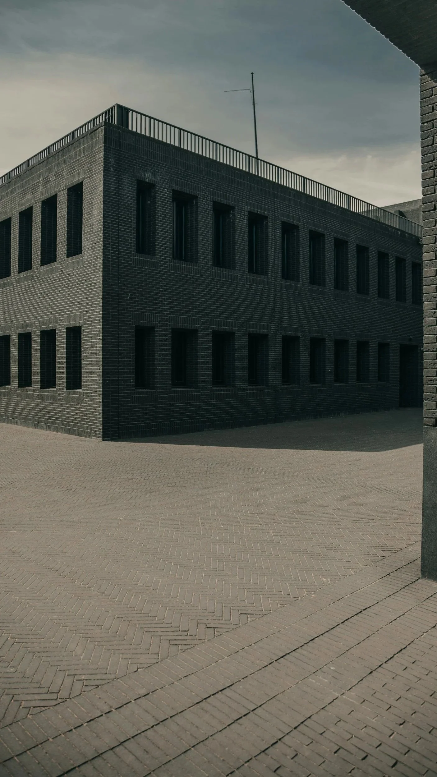 A black brick building with multiple windows and a rooftop antenna, seen from the ground level, with an empty paved area in the foreground and a cloudy sky overhead.