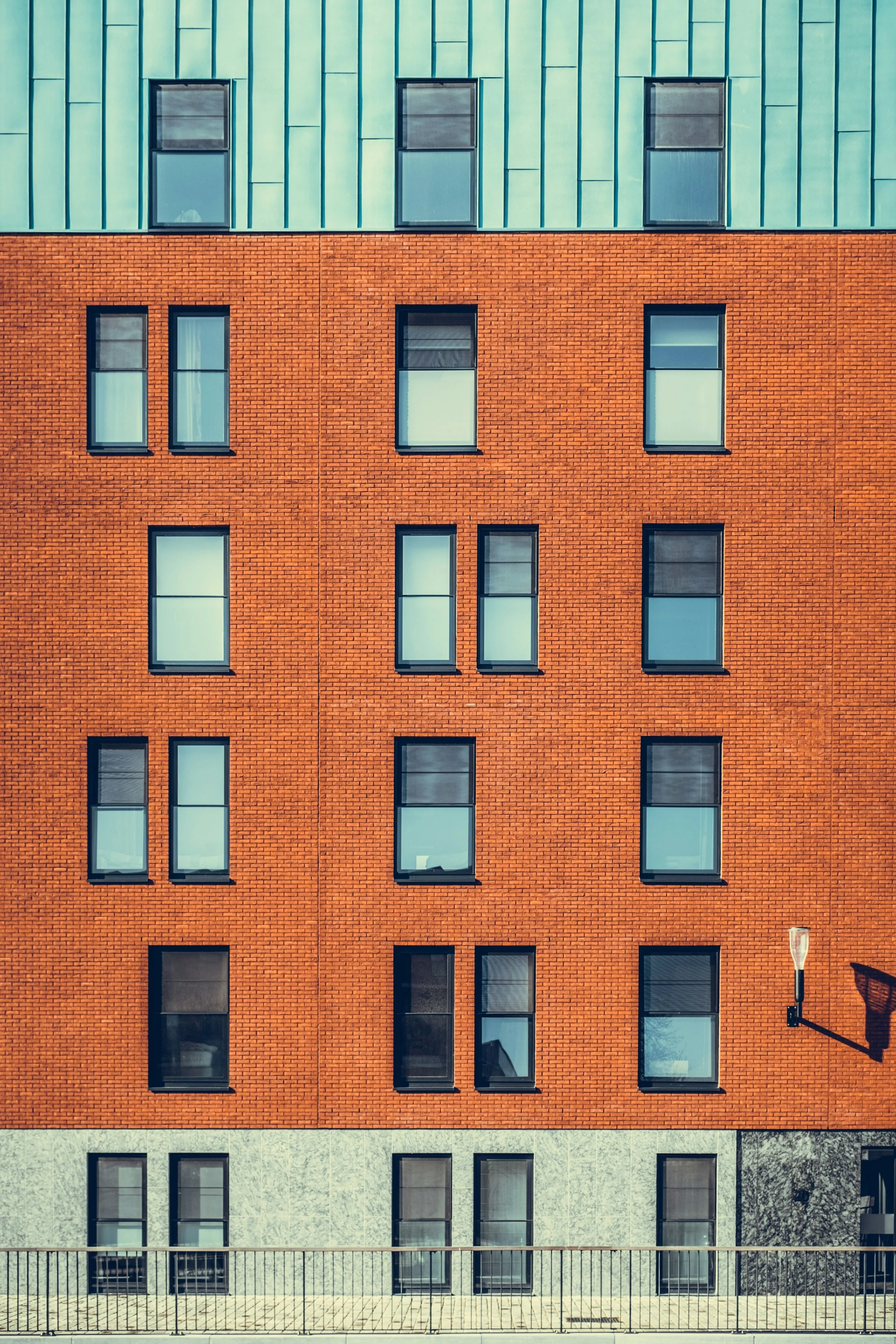 Facade of a modern multi-story building with a brick lower section and a teal upper section, featuring multiple rectangular windows and a street lamp to the right.