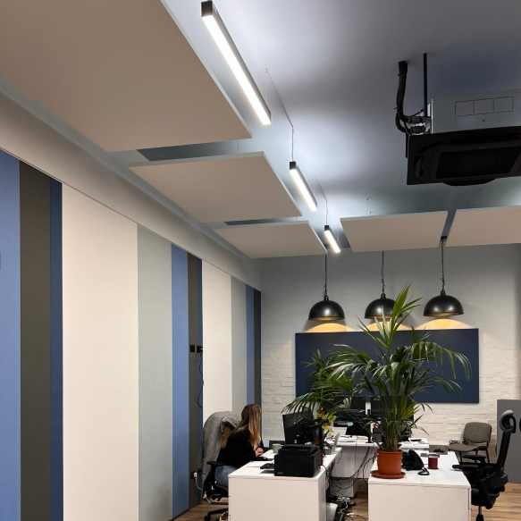 Office interior with white desk, computer, and a woman working, decorated with large green plant and black pendant lights, in a modern workspace.