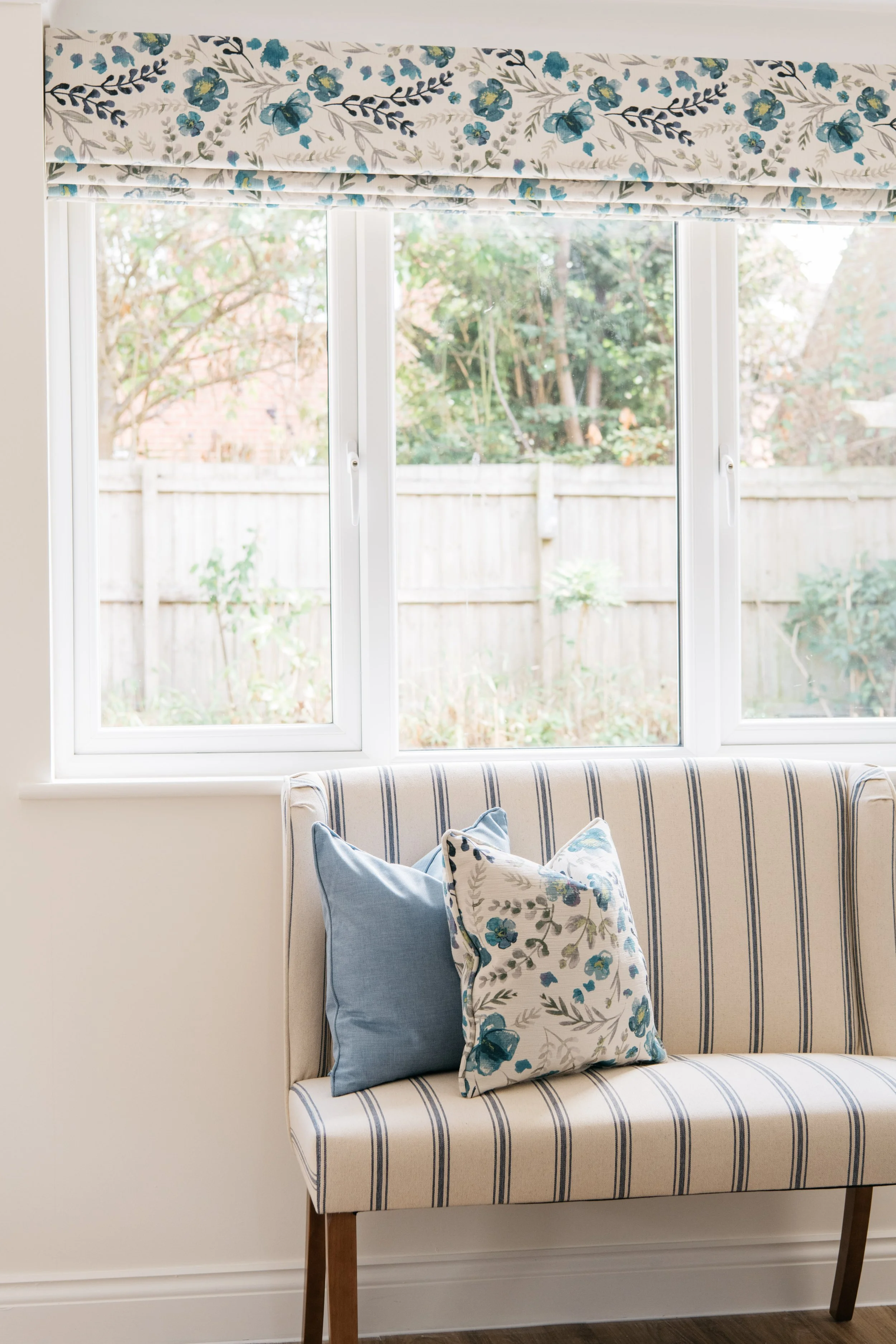 A striped sofa with two blue patterned pillows in front of a window with a bespoke Roman blind and view of a backyard garden.