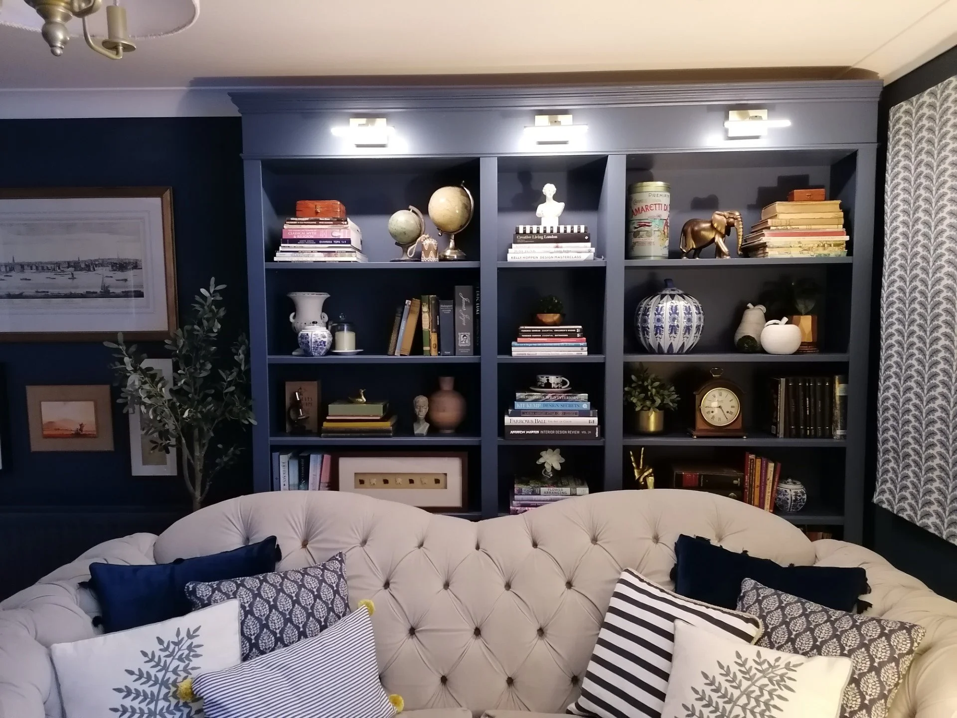 Living room with white tufted sofa, various patterned pillows, dark blue wall with framed artwork, and a dark blue bookshelf filled with decorative objects, books, and globe, illuminated by overhead lighting, with patterned curtains on one side.
