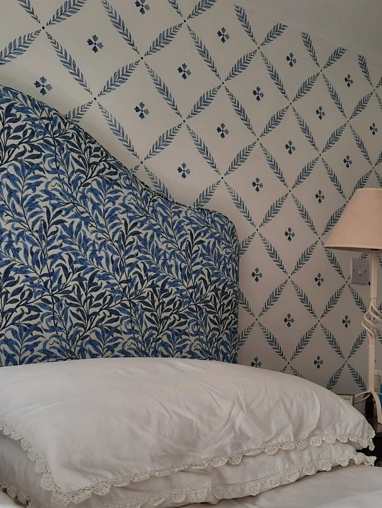 A bed with a white pillow and sheets in front of a bespoke blue and white patterned headboard, with contrasting patterned wallpaper on the wall above.