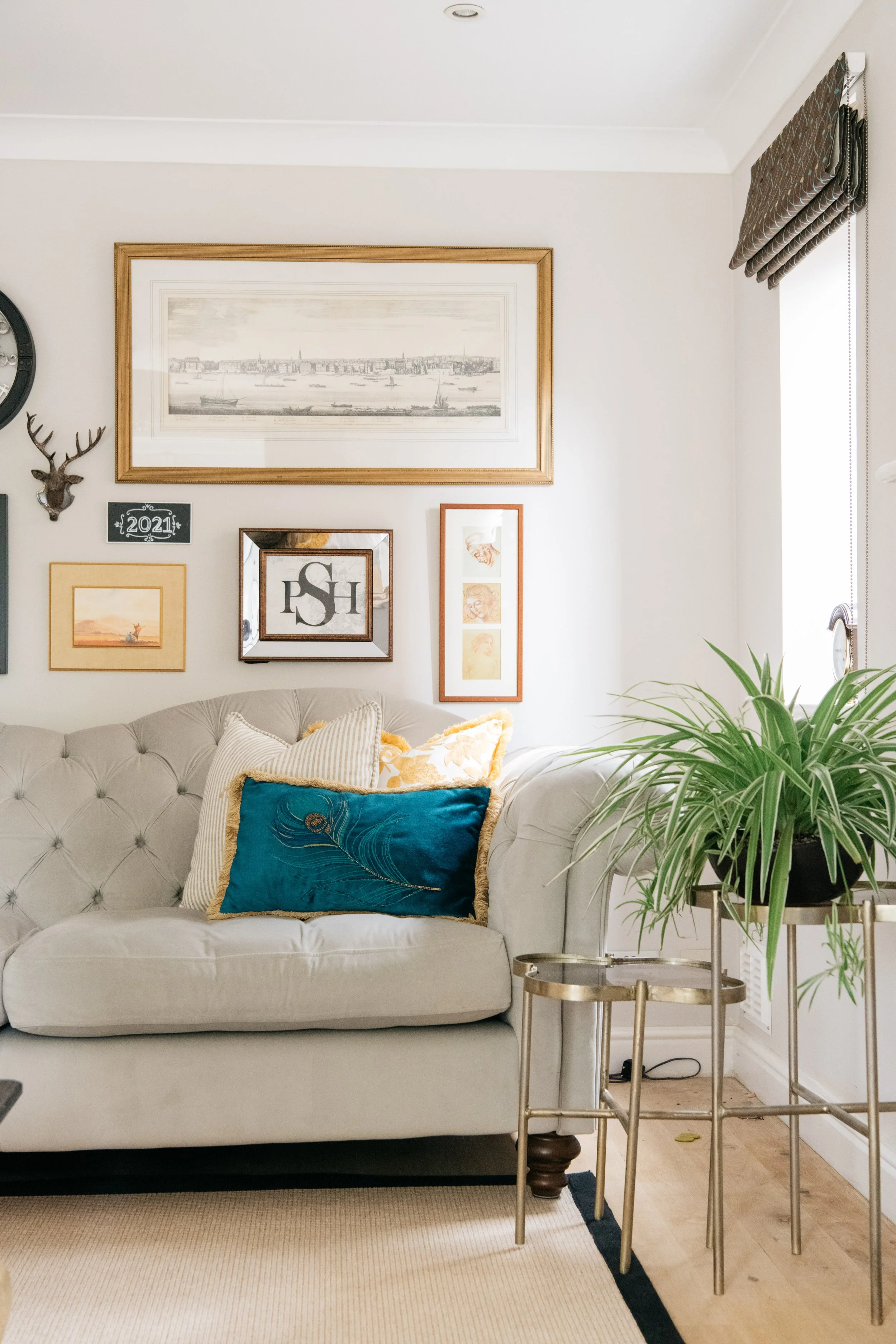 A cozy living room corner featuring a light-colored tufted sofa with beige, white, and teal cushions, a large green potted plant on a gold and glass side table, and a gallery wall with various framed pictures and artwork; sunlight streams through a w