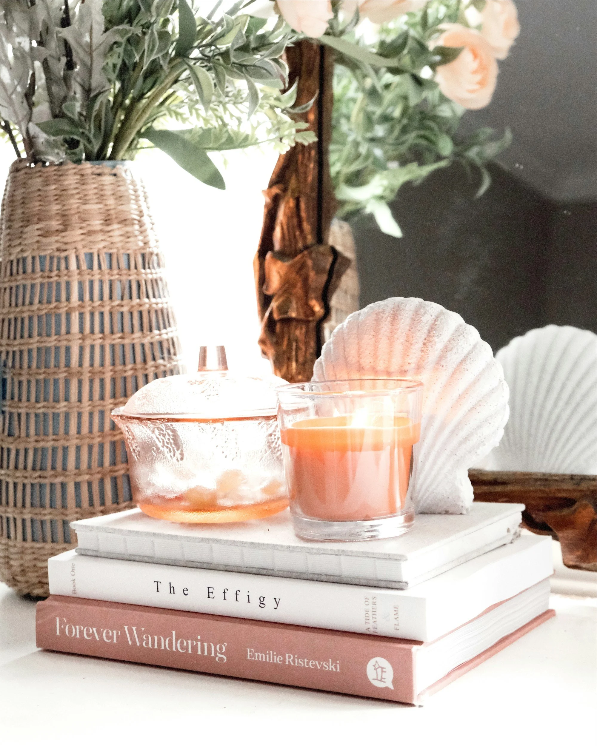 A stack of two books, a pink candle in a glass holder, a decorative seashell, a glass teapot, and a larger seashell, arranged on a white surface with a wicker vase holding green foliage and pink flowers in the background.