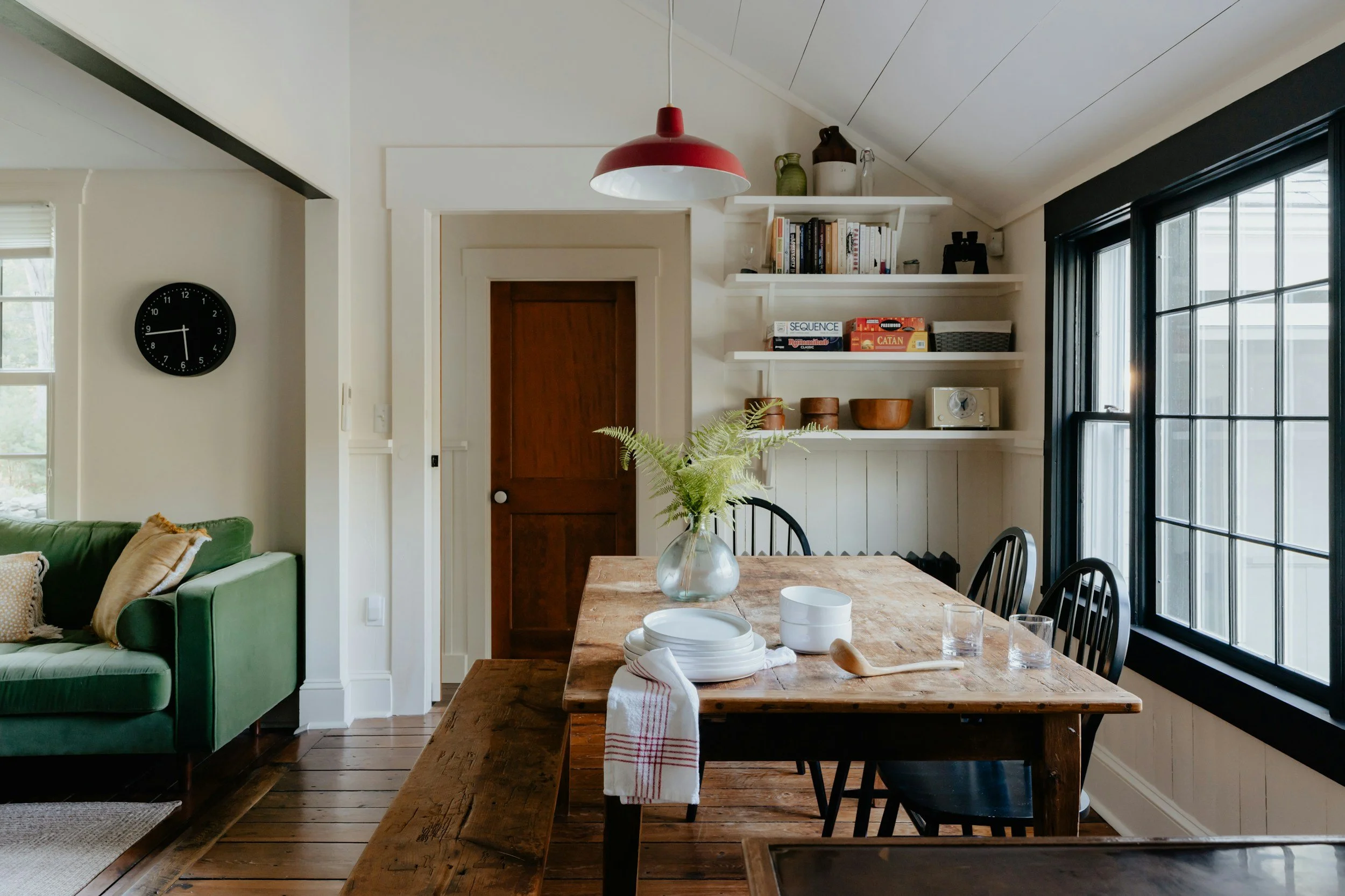A dining area with a rustic wooden table set with white dishes, glasses, and a wooden spoon, decorated with a vase of green fern leaves. There are black chairs around the table, large windows on the right letting in natural light, white shelves with books and decor on the wall, a wooden door in the background, and a black wall clock on the left.
