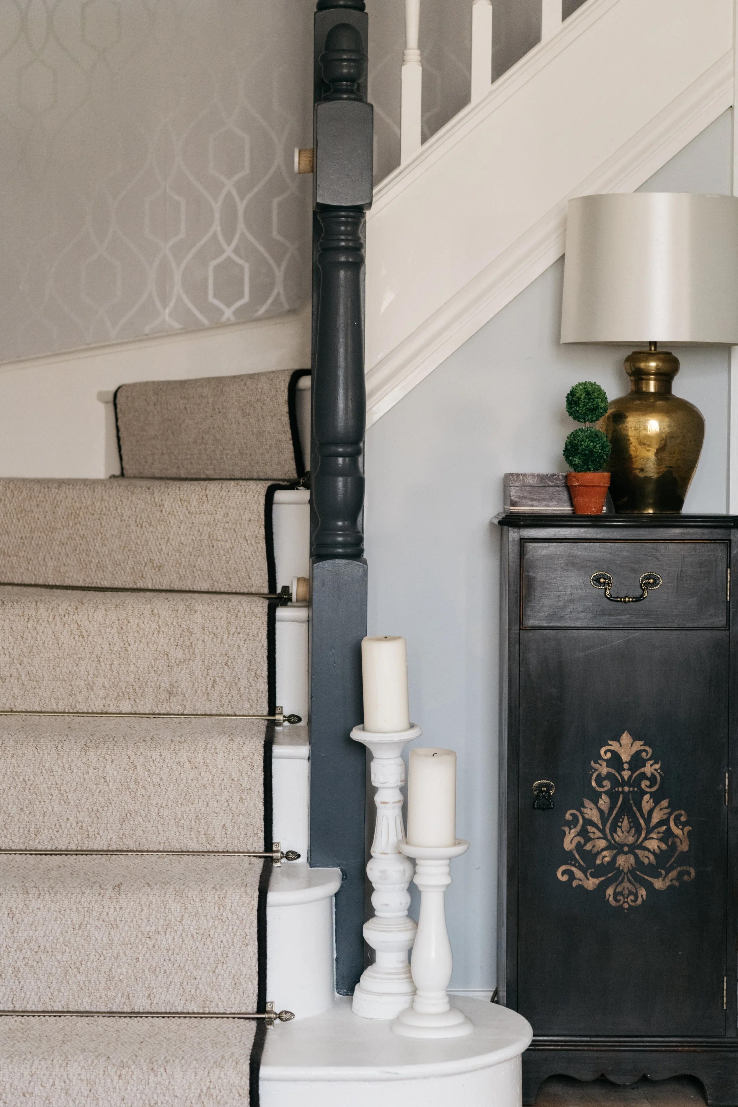Close-up of a staircase with beige carpet, decorated with three white candles on a small white table, next to a dark wooden cabinet with gold decorative details, a large gold table lamp, and two small topiary plants.