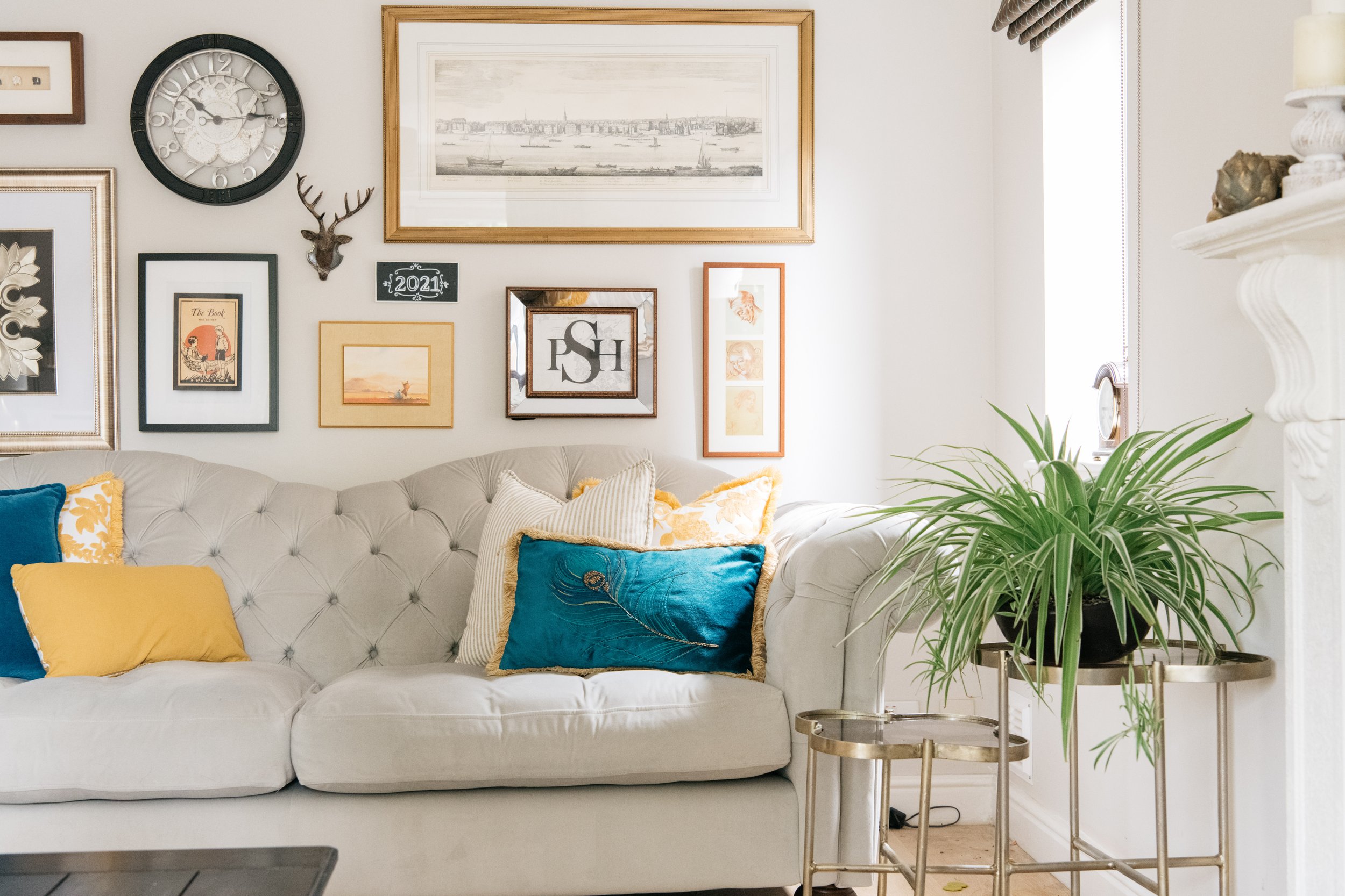 Living room with beige tufted sofa, decorative pillows in blue and yellow, a gallery wall of framed art and a clock, a large green potted plant on a gold stand, and a white fireplace mantel.