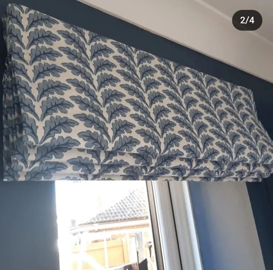 A window with a bespoke blue and white patterned Roman blind featuring leaf designs, partially open, showing a house with a tiled roof outside.