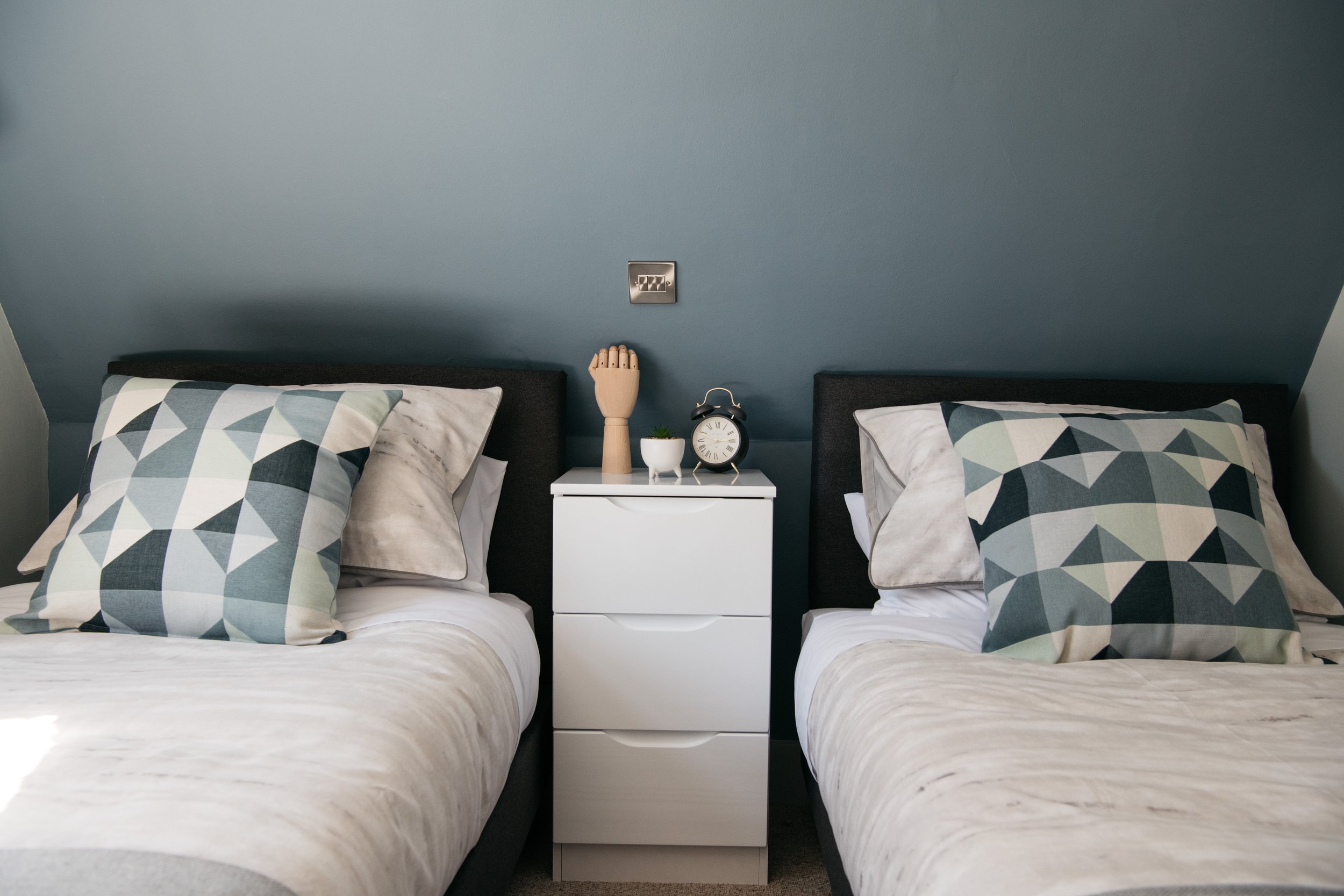 View of two beds separated by a white nightstand, with a wooden hand mannequin, small potted plant, and black alarm clock on top, against a blue wall.